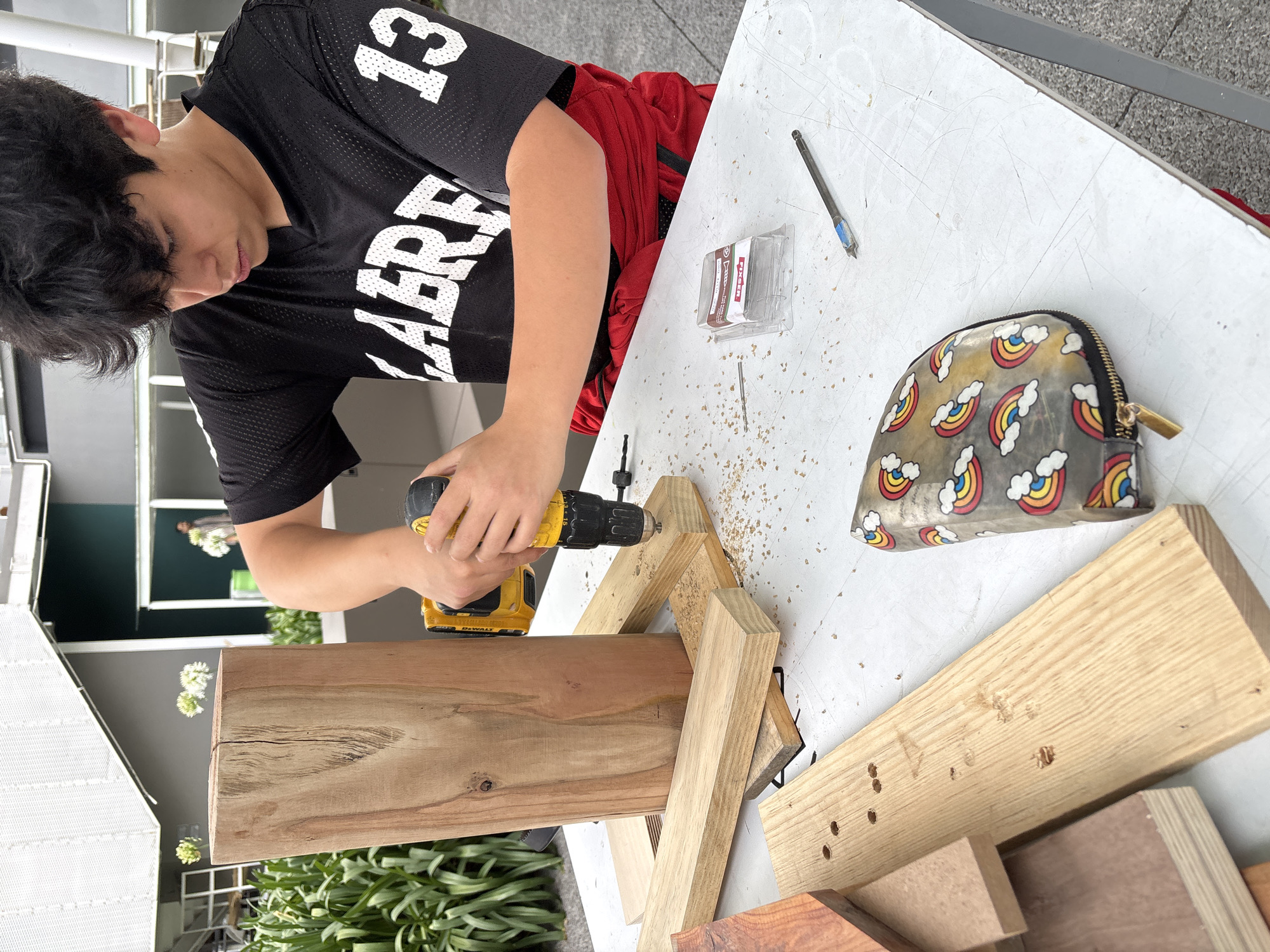 A young person in a black jersey with white lettering is operating a yellow DeWalt drill to assemble wooden components on a white folding table. A package of drill bits, a single drill bit, and a purse with rainbow cloud graphics are also on the table. There is wood shavings from the drilling operation and other pieces of wood present on the table. The location may be outdoors based on the presence of external buildings and landscaping visible in the background. The location in Mexico City, Mexico cannot be independently verified from the image.
