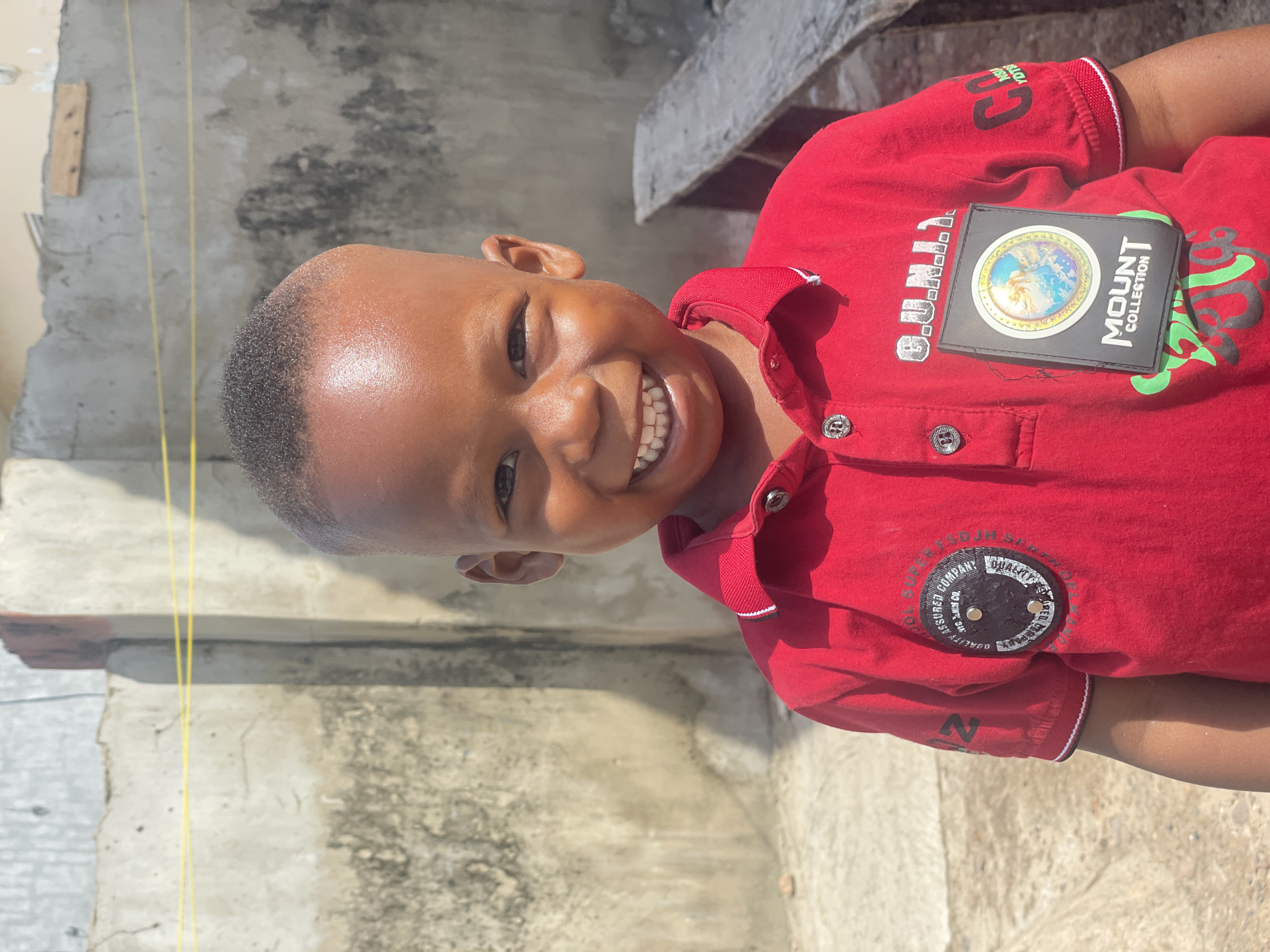 A young boy in a red collared shirt smiles brightly, likely outdoors in Lokoja, Nigeria. The shirt has a "Mount Collection" patch and other designs. The background is a concrete wall with yellow electrical wires.