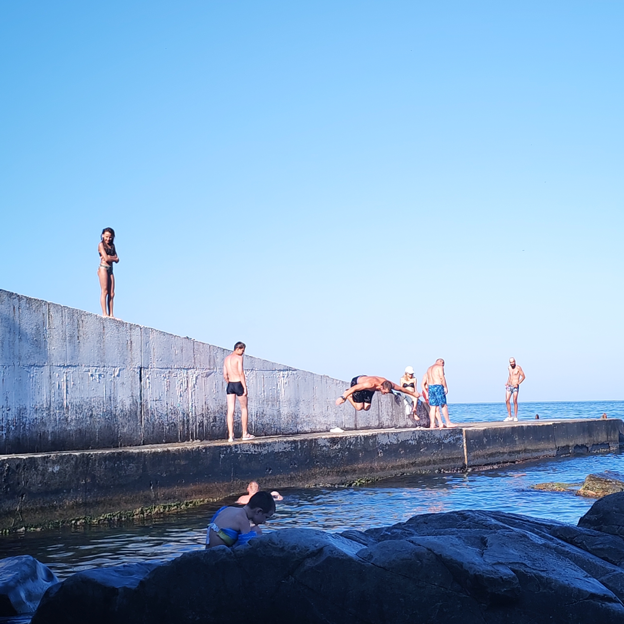 The image depicts a group of people at a concrete embankment by the sea, likely in Partenit, Ukraine, on a clear day with a bright blue sky. The embankment is wide and appears to serve as a pier or a breakwater. On the far left, a young woman in a bikini stands on top of the embankment, arms crossed, looking towards the viewer. To her right, a man in black swim trunks stands on the embankment, facing away from the camera. Further to the right, a man is captured mid-air, performing a dive into the water. Behind him, a woman in a white hat and bikini and another person are standing, partially obscured. To the far right, a man in patterned swim trunks stands facing the sea. In the water below the embankment, several people are visible. A young boy wearing arm floaties is in the foreground, looking down into the water near some dark rocks. Another person is partially submerged in the water further out. The sea is calm and blue, with some small waves. The concrete embankment shows signs of wear and tear, with some green algae visible along its lower edge.