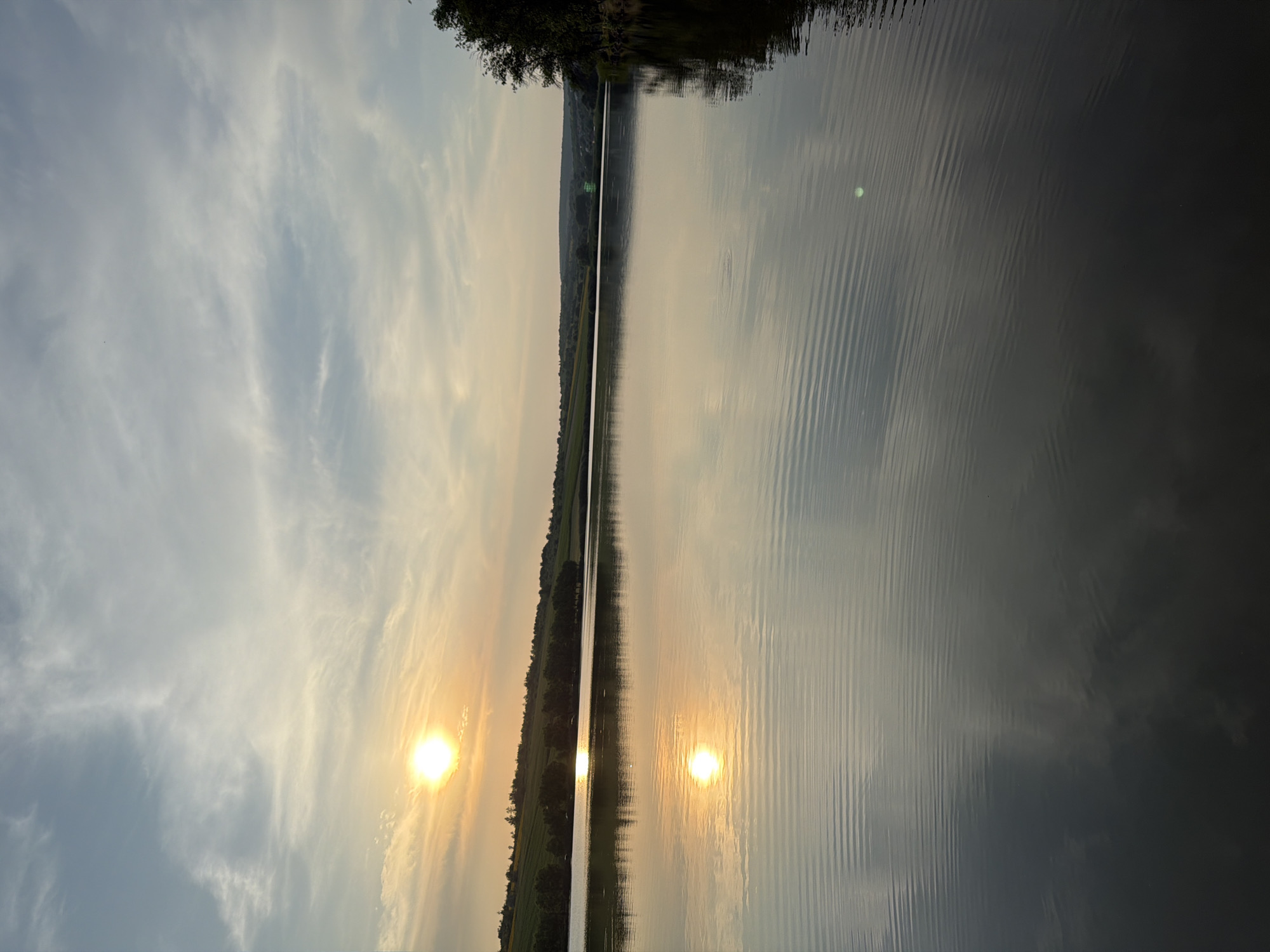 The image depicts a tranquil lake scene at sunset. The sun, a bright orb tinged with yellow and orange, is low in the sky, casting a warm glow. Wispy clouds, ranging in color from white to light grey, are scattered across a pale blue sky. The sun's reflection is clearly visible on the calm surface of the lake, creating a mirror image. The shoreline in the background is lined with a dense row of trees, behind which rolling hills are visible, some covered in greenery or agricultural fields. The overall atmosphere is serene and natural. The location is identified as Găleștii Noi, Moldova. There are no people or specific objects of interest other than the natural landscape.