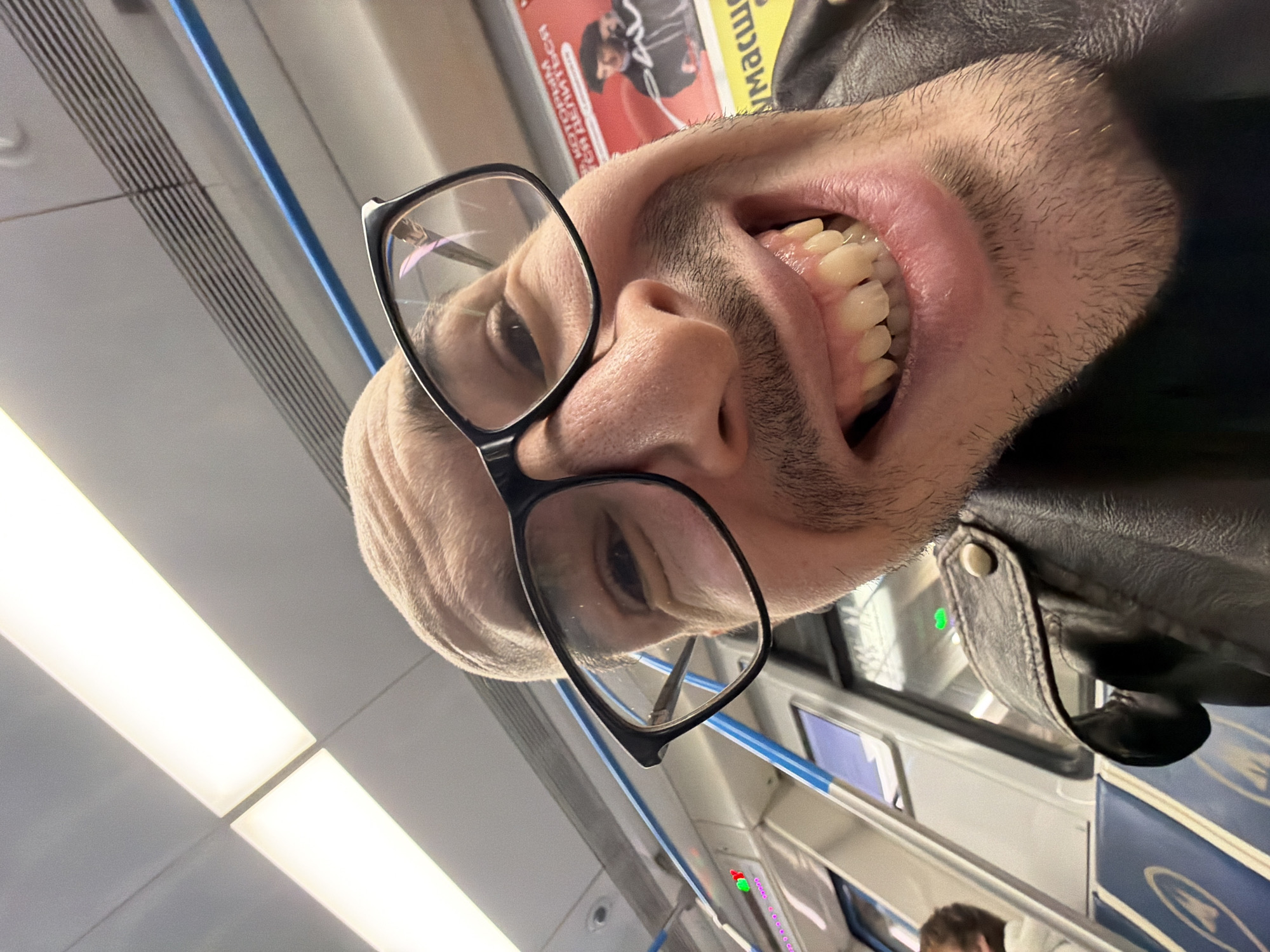 A photograph depicts a man wearing eyeglasses and a black leather jacket inside a train in Moscow, Russia. The image is rotated, showing the man's face in close-up from a low angle. His mouth is open, revealing his teeth. The background shows the interior of the train car, including seating and informational panels. The location is indicated by the train's interior design and signage, consistent with Moscow metro trains.