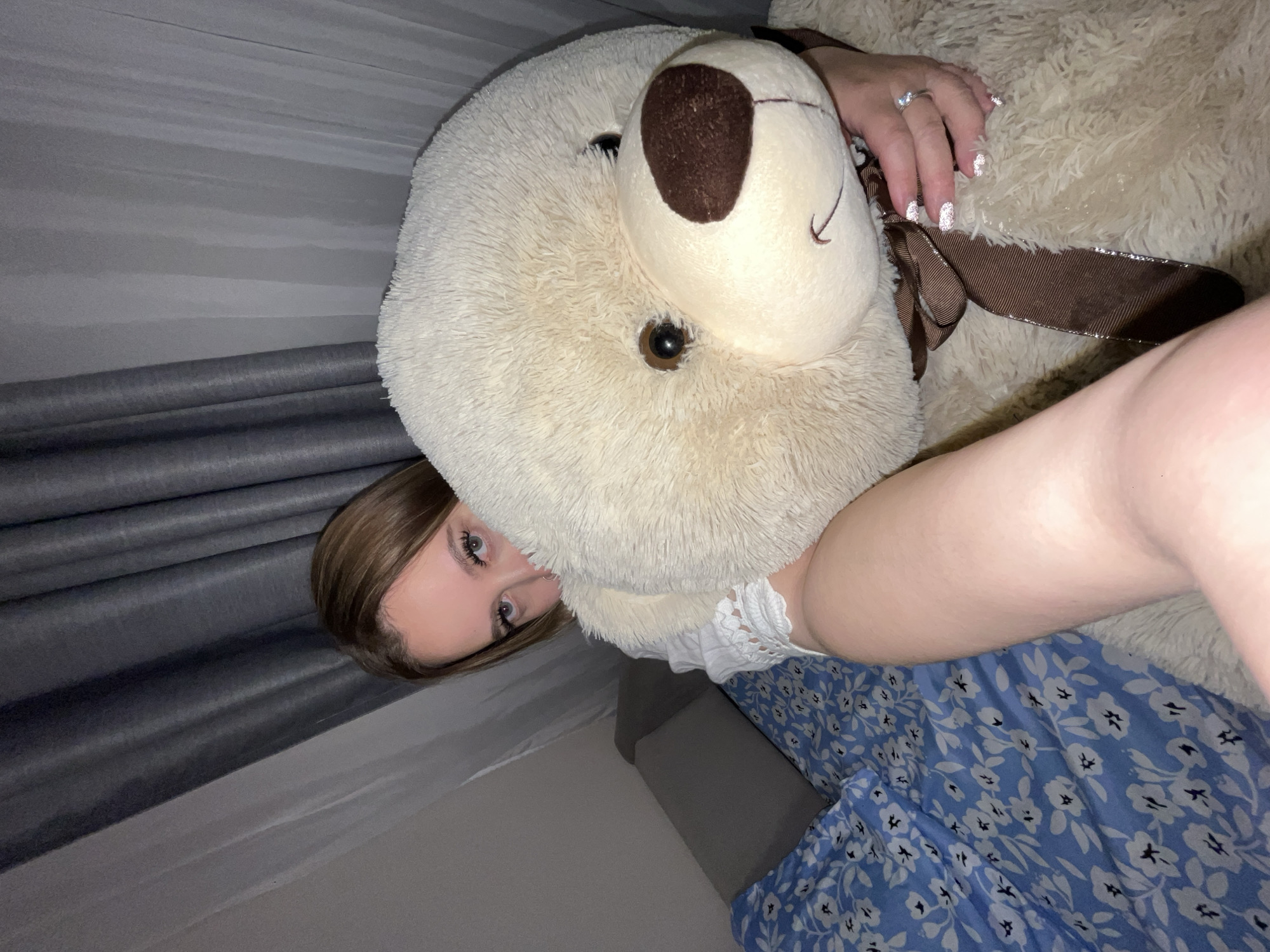An indoor scene in Krasnodar, Russia, captures a young woman embracing a large, cream-colored teddy bear. The woman's face is partially visible, showing her light-colored eyes and light brown hair as she looks towards the viewer. She is wearing a white top with lace detailing and a blue skirt or dress adorned with a white floral pattern. Her left hand, featuring glittery nail polish and a ring on her ring finger, gently rests on the teddy bear's fluffy head.
The oversized teddy bear has dark brown eyes and a matching brown nose, with a brown ribbon tied around its neck. The background suggests a cozy setting, possibly a bedroom or living room, with a grey striped wall or headboard visible. Dark grey rolled fabric, possibly curtains or another textile, can be seen on the left side of the frame. The lighting indicates it might be evening or an artificially lit environment, contributing to a soft ambiance. The overall impression is one of comfort and relaxation. No visible text is present in the image.
