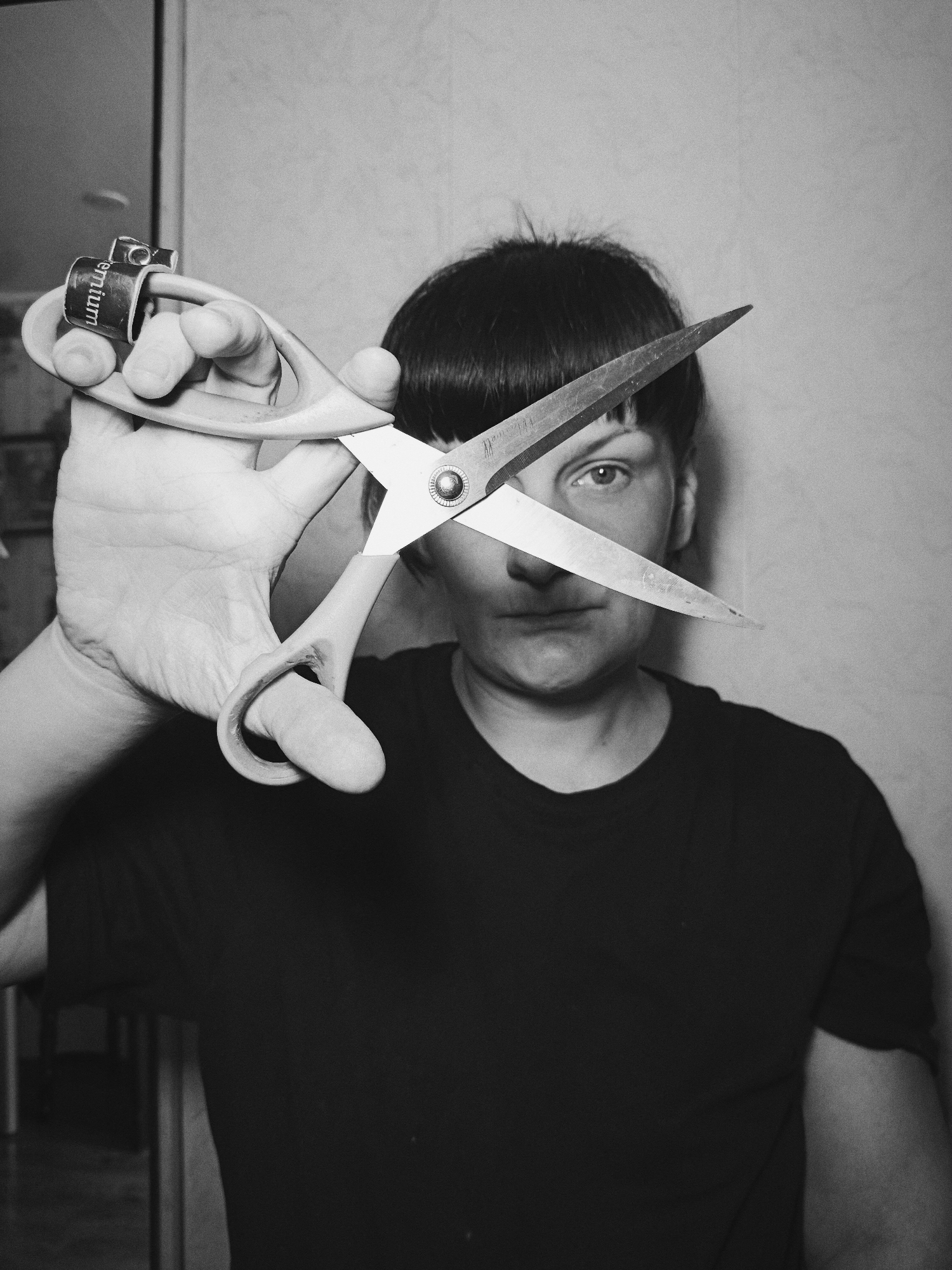 The image is a black and white photograph of a person holding a pair of open scissors up to their face. The blades of the scissors bisect the face horizontally, positioned between the nose and eyes. The person has dark hair and is wearing a dark t-shirt. The background appears to be a plain wall. The brand name on the scissor handle reads 'Premium'. The image location is unknown, despite the prompt mentioning Фоки, Russia.
