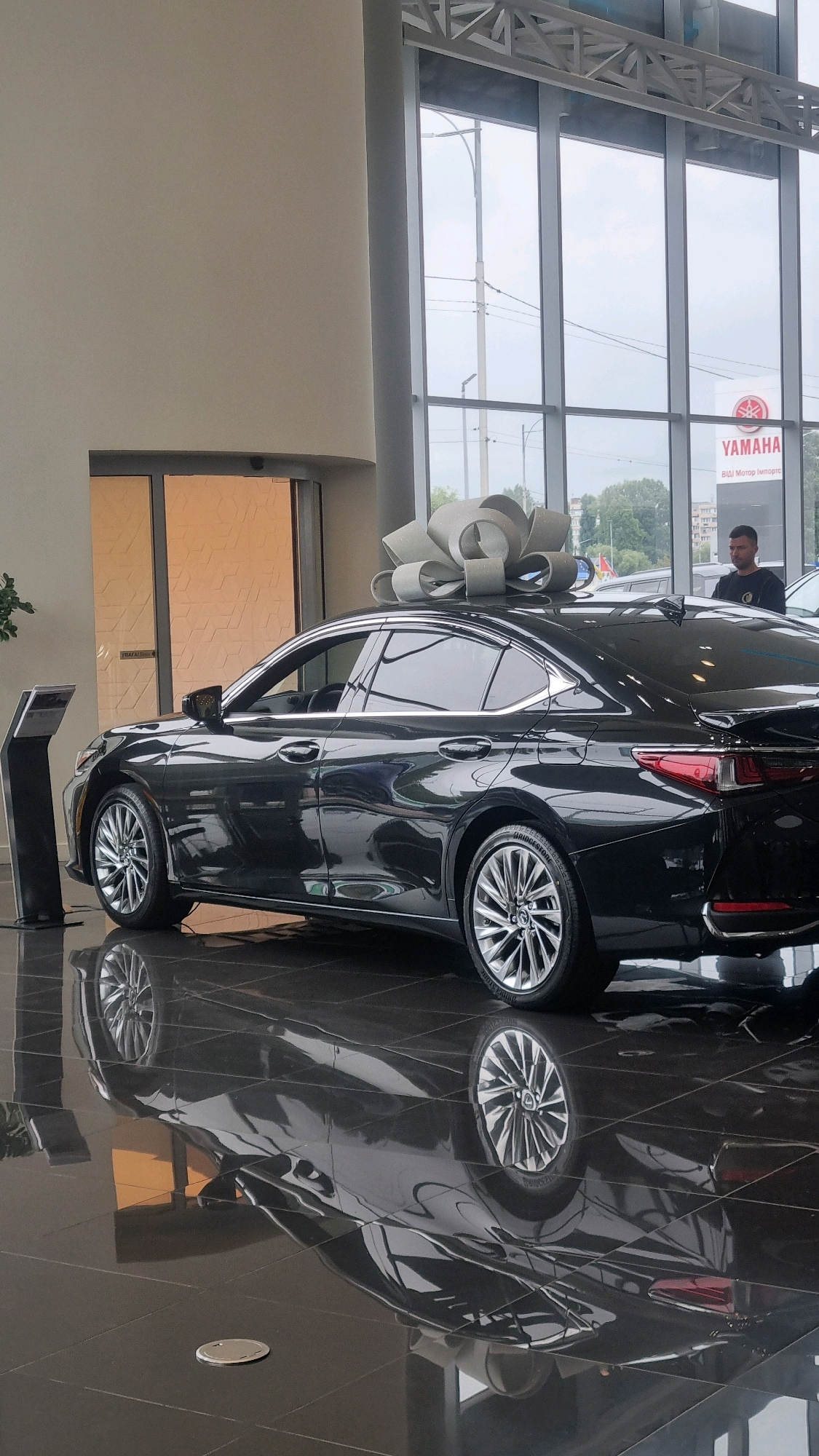 A black Lexus sedan, adorned with a large grey bow on its roof, is displayed inside a modern car dealership in Sofiivska Borshchahivka, Ukraine. The car is positioned on a highly reflective, dark floor, which mirrors its shape. Behind the car, large windows reveal an overcast sky, trees, and a streetlamp. A sign for "YAMAHA" is visible through the windows. A man in a dark t-shirt stands to the right of the car, looking towards the vehicle. To the left of the car, a black information kiosk stands on a dark tiled floor. The dealership interior features light-colored walls and a grey structural ceiling.