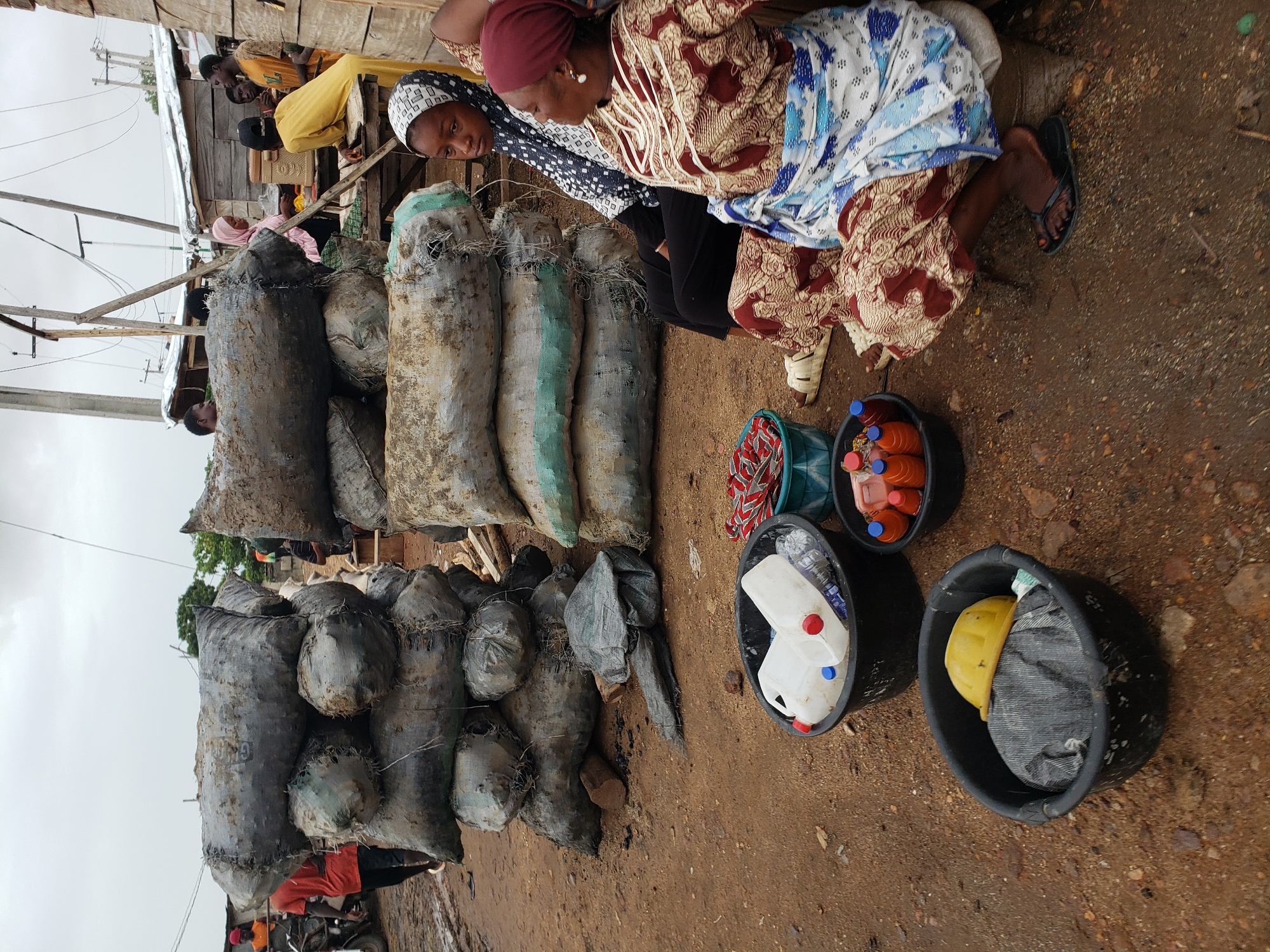 This image depicts an outdoor market scene in Lokoja, Nigeria. The focal point is a roadside stall with piles of large, heavy sacks, likely containing goods for sale. Two women, dressed in traditional African clothing, are kneeling beside the sacks, seemingly attending to their wares. Beside them are several black buckets containing various items, including bottles of liquid and plastic containers. The ground is dirt, showing signs of recent rain. In the background, there are glimpses of wooden structures and other people, indicating a bustling market environment. The overall atmosphere is one of activity and commerce.