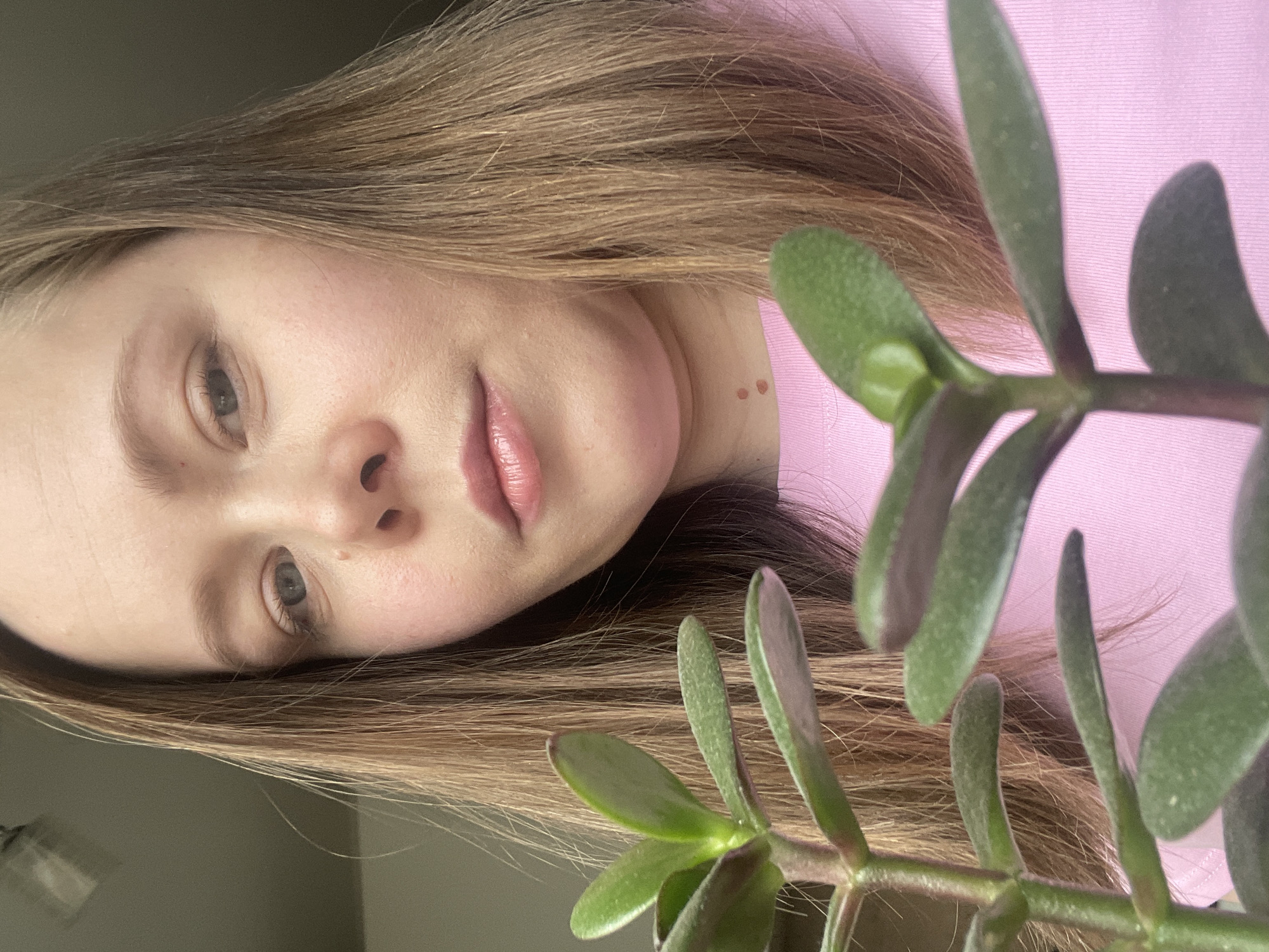 A woman with light brown hair and fair skin is featured in the image. Her face is angled towards the camera, and she is wearing a pink shirt. A plant with green leaves is positioned in the foreground, partially obscuring her. The background is a light, neutral color. The image's location is unspecified and cannot be determined to be the city of Chaikovsky, Russia, based on the visual information provided.