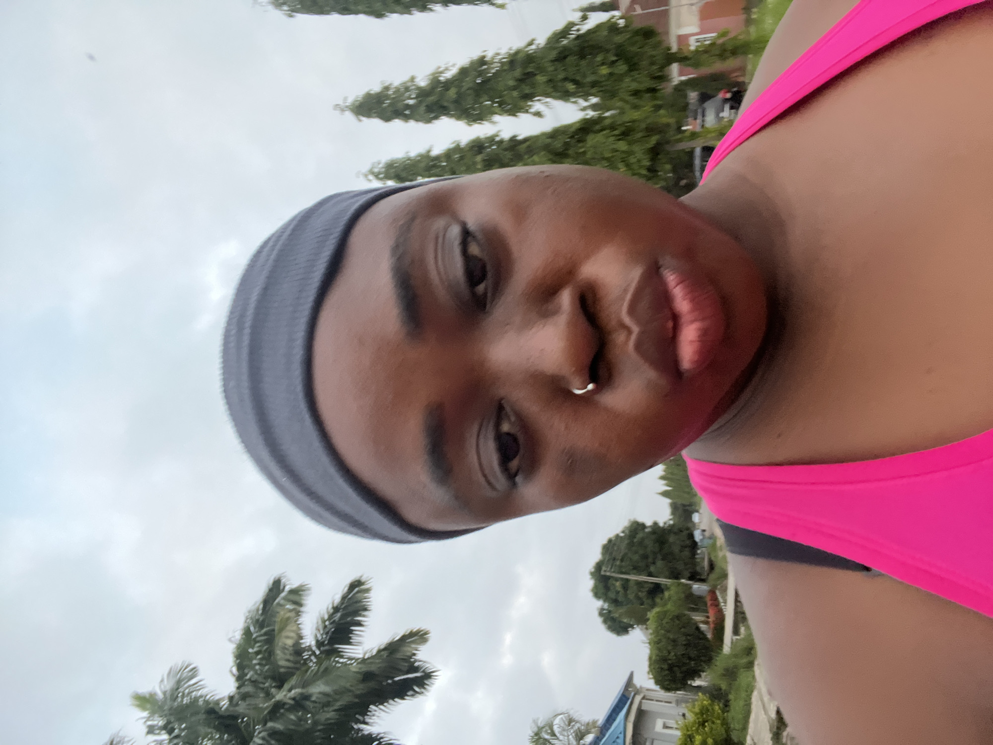 A medium shot shows a woman with dark skin, a grey headband, and a bright pink tank top from the chest up, looking forward with pursed lips. She has a nose ring. The background is an outdoor scene with palm trees, tall green trees, houses, and a cloudy sky. The overall impression is a selfie taken outdoors in a residential area. The specific location within Abuja, Nigeria is not identifiable from the visual information provided.