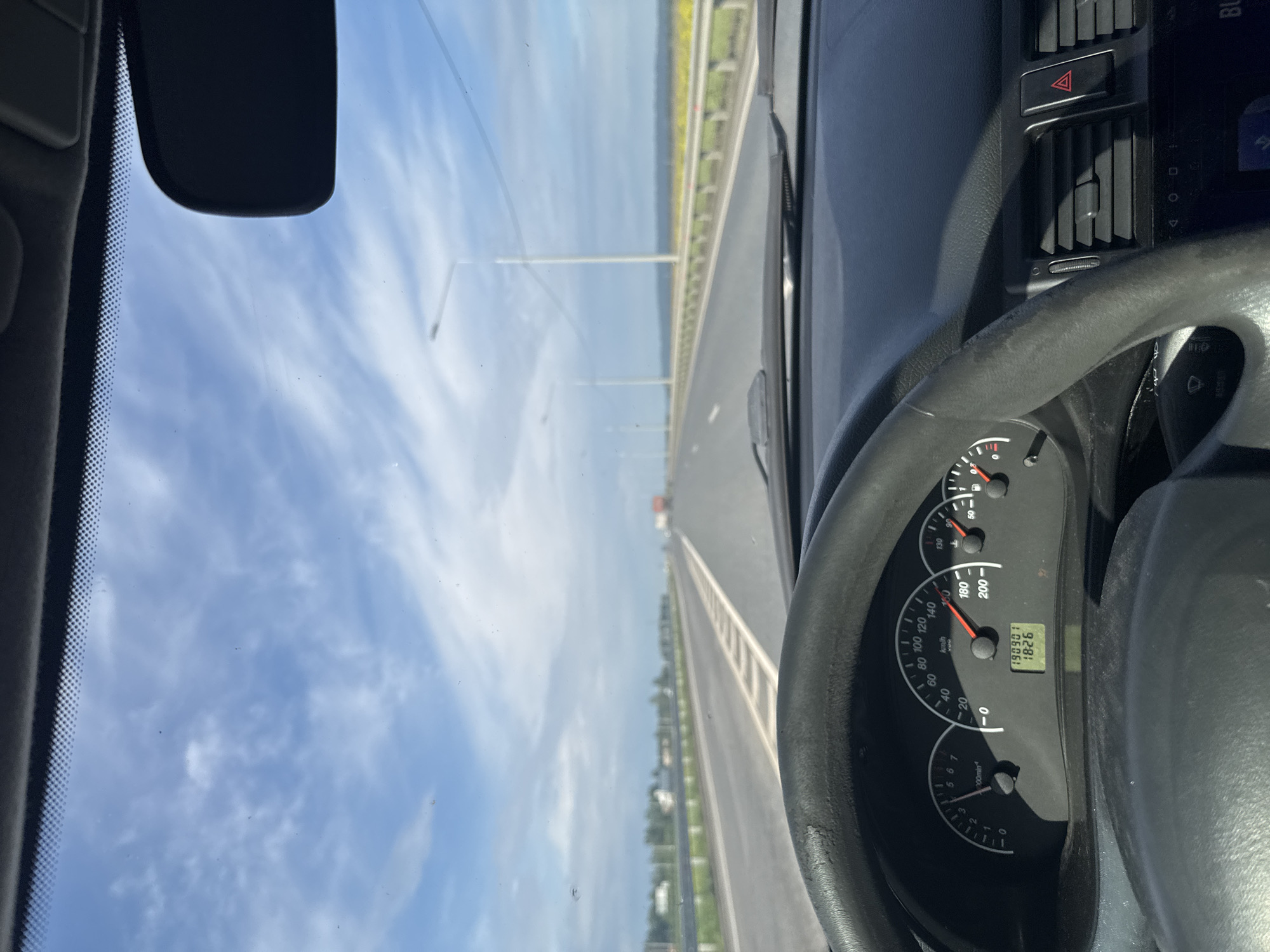 The image presents a view from inside a car, rotated 90 degrees clockwise from a standard forward perspective. The right side of the frame is occupied by the car's dark interior, including a portion of the steering wheel and the instrument cluster. The left side features a bright blue sky with scattered white, wispy clouds, seen through the windshield.
The main subject is a long, straight road extending vertically into the distance, visible through the windshield. The road appears to be a highway or a bridge, with a grey guardrail running along its left side. Multiple white light poles line the road, receding towards the horizon. A small, distant vehicle, possibly red, can be seen far ahead on the road. Beyond the guardrail, a flat landscape, possibly fields or water, stretches towards the distant horizon. The overall setting suggests travel on an open road in Усть-Сыны, Russia, likely near the Krasnoyarsk Reservoir.
The car is in motion, as indicated by the speedometer. Visible details on the dashboard include a black steering wheel with a textured grip, and an instrument cluster featuring a speedometer, tachometer, and digital display. The speedometer needle points to approximately 80 km/h. The digital display shows "190901" (likely odometer or trip mileage) and "18:26", indicating the time is 6:26 PM. The weather is clear and sunny. A red triangular hazard light button and air vents are also visible on the dashboard.