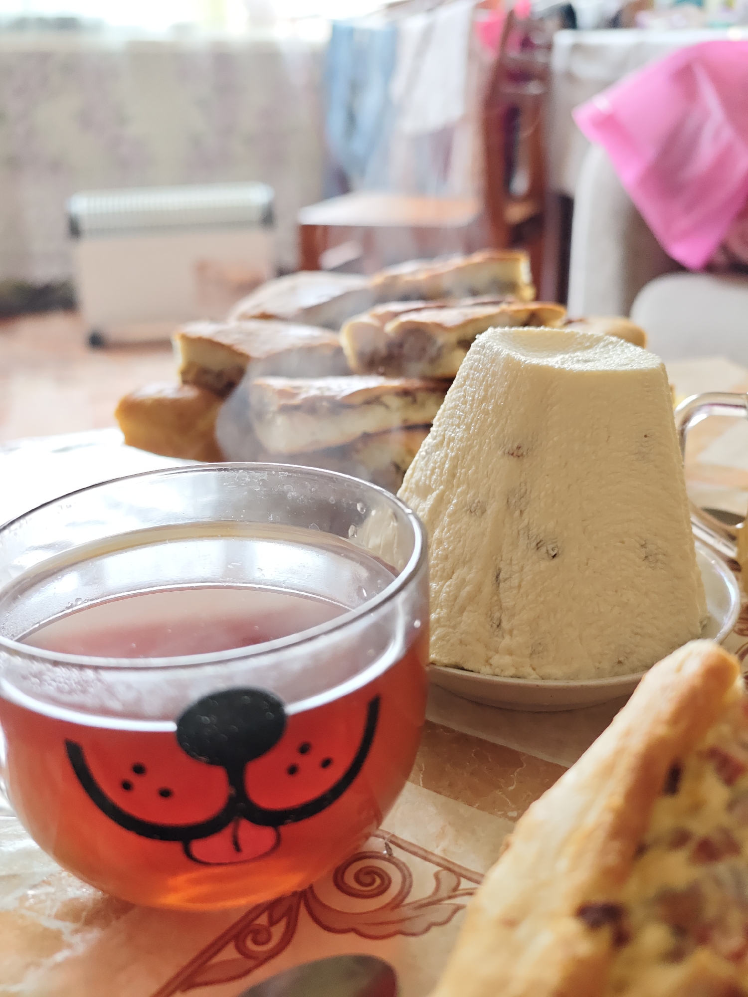 The image shows a table setting with various food items and a cup of tea. In the foreground, a clear glass cup with a cartoon dog face design contains tea. Next to it, on a plate, is a cone-shaped white dessert, possibly paska, with dark specks. Behind these items, there's a stack of sliced pastries, and steam is visible, suggesting they are freshly baked or warm. A piece of savory pie is visible on the right. The background shows a blurred interior with a heater, chairs, and other furniture, indicating an indoor setting. The location is Уральское, Russia.