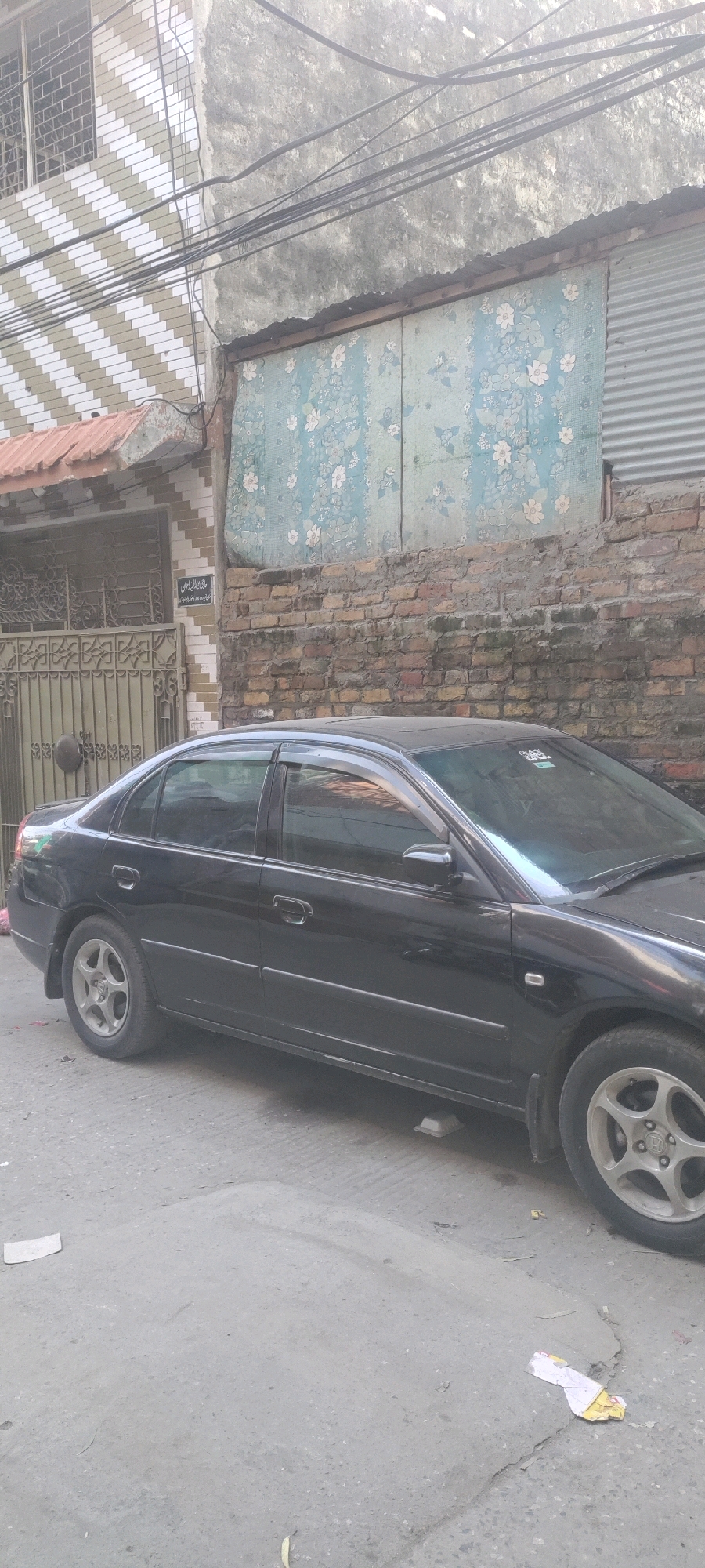 A black sedan is parked on a paved street in front of a building in Rawalpindi, Pakistan. The building has a brick facade, a patterned wall with blue and white flowers, and a metal gate. Overhead, electrical wires are strung across the scene. The car's windows are tinted, and its tires are visible. The street is worn and shows signs of age and use. The overall impression is one of a typical urban street scene in a Pakistani city.