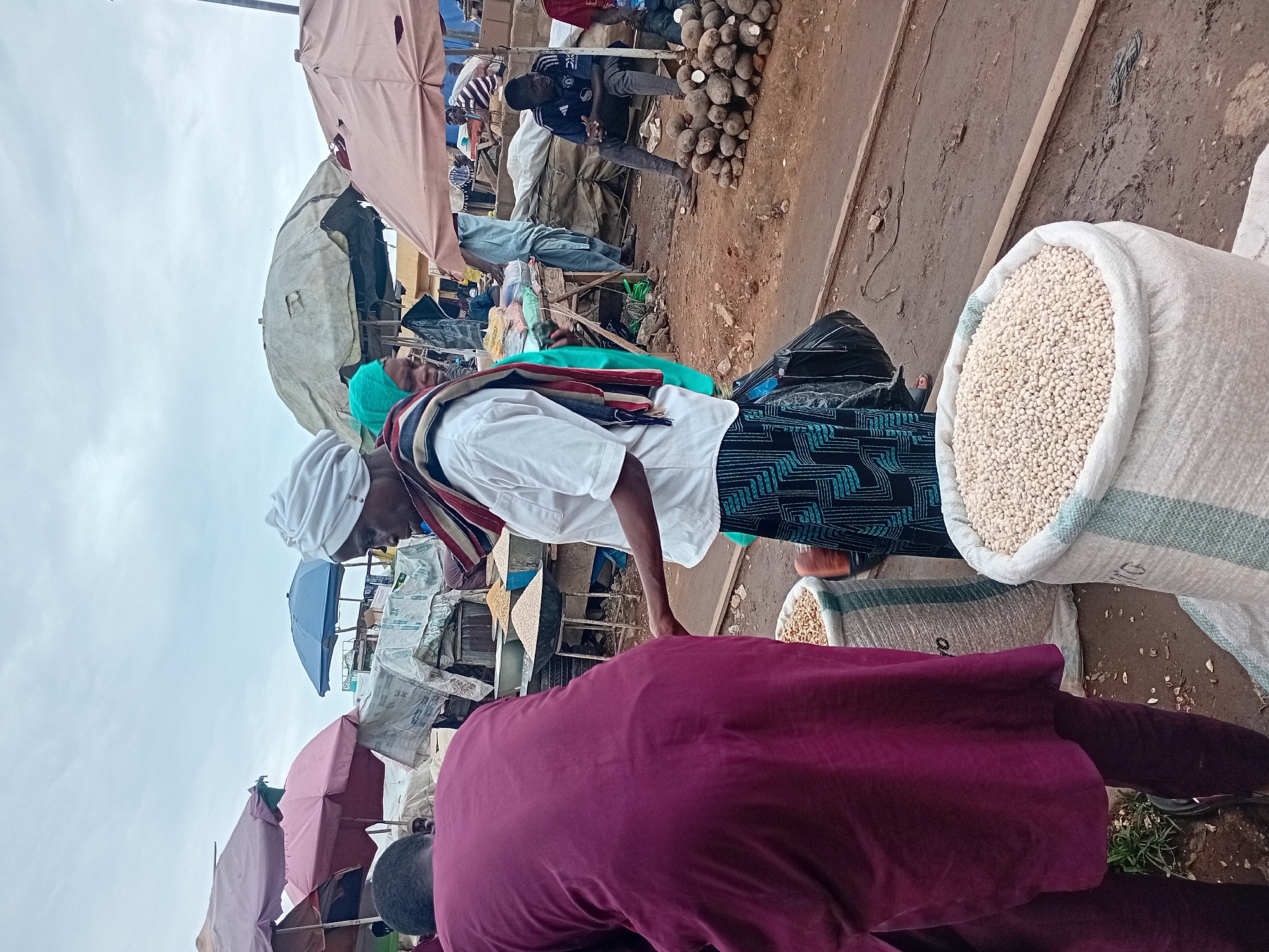 The image depicts an outdoor market scene in Maiburiji, Nigeria. In the foreground, two large sacks filled with white beans are prominently displayed. A person wearing a deep purple garment is partially visible, facing away from the camera and towards the sacks. To the right, another person wearing a white shirt and a patterned skirt is interacting with the sacks, possibly examining or weighing the beans. In the background, various market stalls are set up under colorful umbrellas. Several people are seen browsing or working in the market. The ground is dirt, and the sky is overcast. The overall atmosphere suggests a bustling commercial environment.