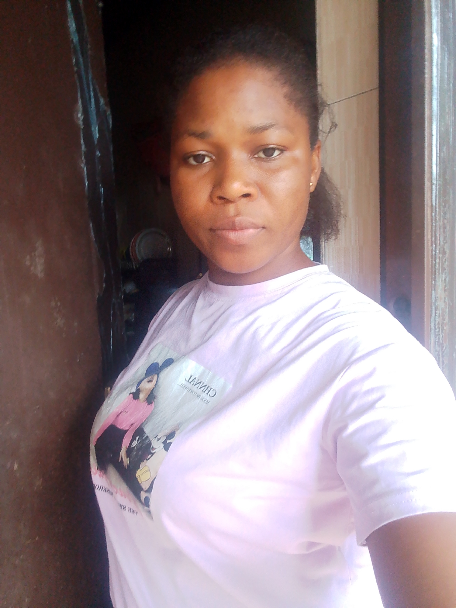 This is a medium shot photograph of a young woman with brown skin and dark hair, looking directly at the camera. She is wearing a light pink t-shirt with a graphic print on the left side of her chest. The graphic appears to be a fashion illustration of a woman in a pink top and black skirt, alongside cartoon characters. Some text is visible on the t-shirt, appearing to be reversed or mirrored, possibly "CHINVA" or a similar word.
The woman is standing in what appears to be an indoor setting with a slightly cluttered background. To her left is a dark, textured surface, possibly a wooden door or wall. To her right, a lighter colored wall with vertical lines is visible, suggestive of wooden paneling or blinds, with a bright light source to the far right, indicating an opening like a window or doorway. In the background, on the left side, some stacked plates and other items are partially visible, suggesting a domestic environment.
The lighting suggests it is daytime, with natural light illuminating the scene from the right. The woman's expression is neutral and direct. The overall impression is that of a casual, candid photograph taken indoors, possibly in Yenegoa, Nigeria, given the provided location context.