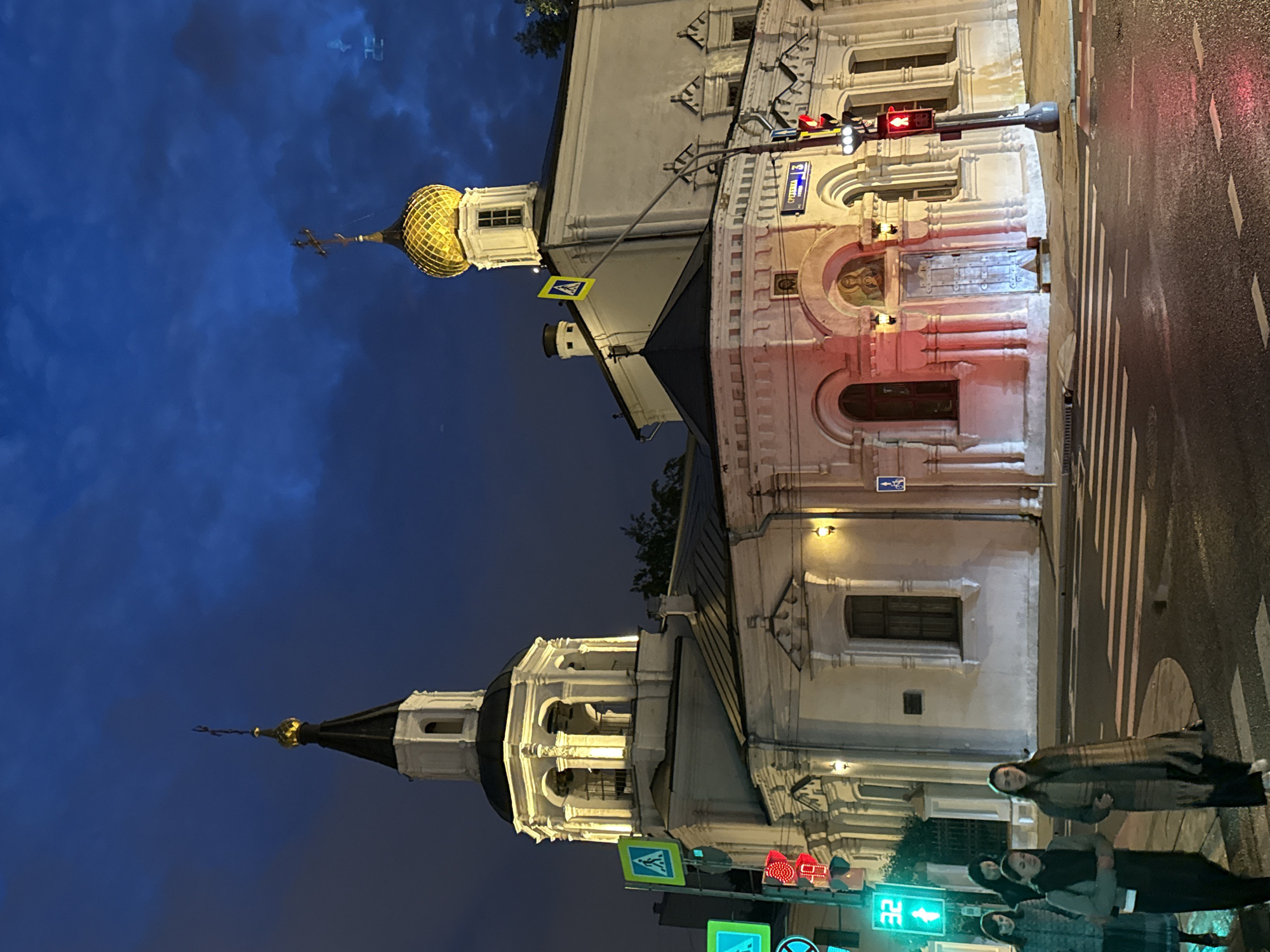 The image captures a night scene in Moscow, Russia, showcasing the architecture of Russian Orthodox churches against a dark blue sky. One church features a golden onion dome, while the other has a black-topped spire. Streetlights illuminate the buildings, emphasizing architectural details like arches and facades. In the foreground, pedestrians are seen crossing a street with crosswalks. Traffic lights are visible, indicating the status of the intersection. The street appears wet, possibly from recent rain, reflecting the surrounding lights.