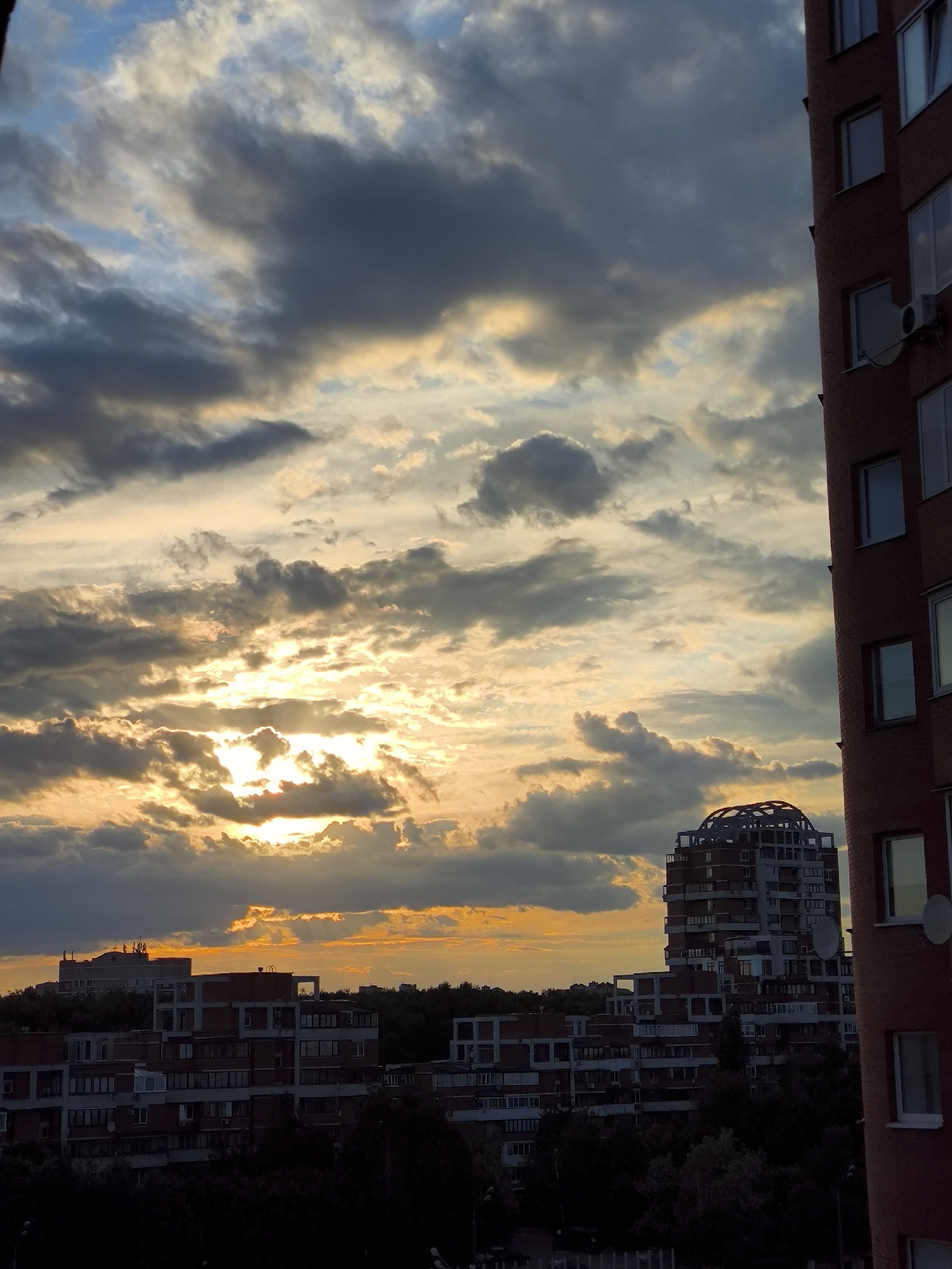 The image depicts a dramatic sunset or sunrise over an urban or suburban landscape, viewed from an elevated perspective. The sky dominates the upper two-thirds of the frame, filled with a dynamic mix of dark, heavy clouds and brightly illuminated, golden-orange clouds. The sun is partially obscured by these clouds in the center-left, creating intense rays of light that pierce through, casting a warm glow across the sky.
On the far right, a tall, brick-colored building with multiple windows and an air conditioning unit is partially visible, acting as a foreground element. In the mid-ground, a distinctive multi-story building with a unique, rounded, lattice-like dome structure on its roof stands out. Below and beyond these, a collection of other residential or commercial buildings, appearing darker due to the backlighting, stretch across the lower half of the image. The silhouettes of trees and vegetation are visible beneath the buildings.
No people or explicit activities are visible, making the dramatic sky and urban architecture the primary subjects. The time of day is clearly either sunset or sunrise, evidenced by the warm, intense light and the contrast between the bright sky and the increasingly shadowed ground features. There is no visible text. The scene conveys a sense of natural beauty and urban life converging at a transitional moment of the day.