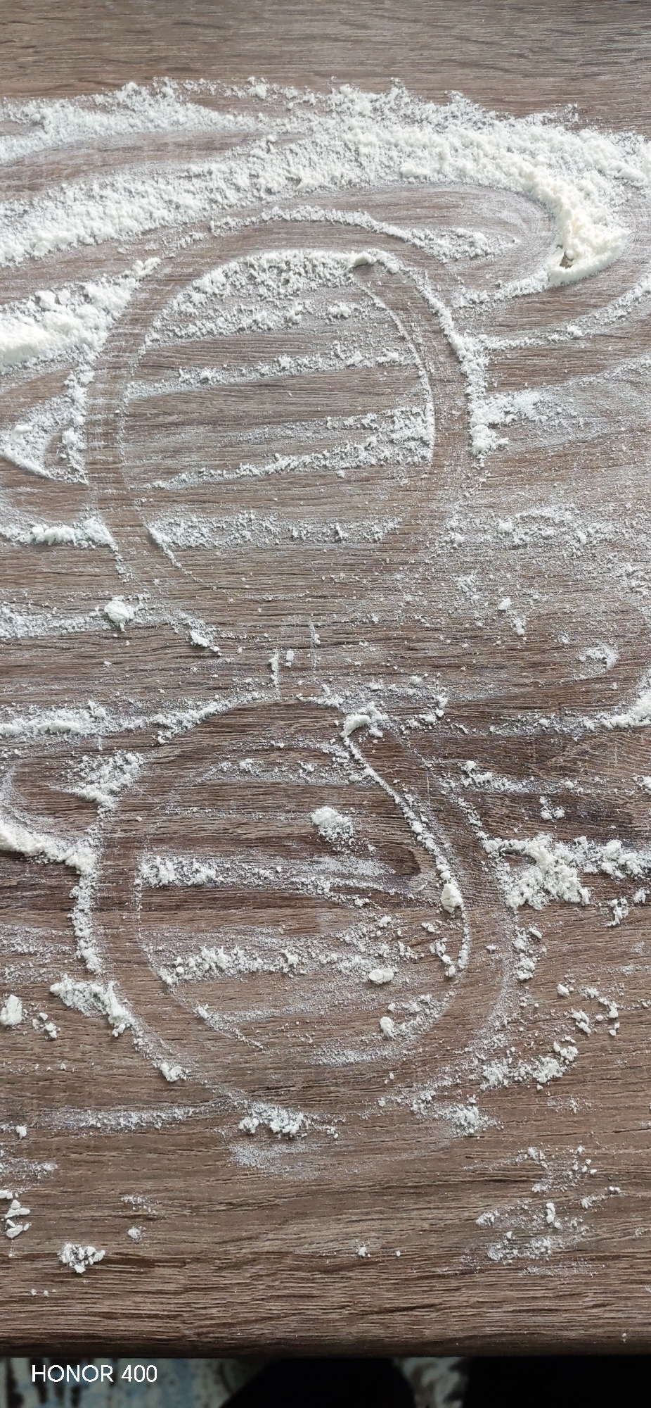 The image displays a close-up, overhead view of a wooden surface, likely a kitchen countertop or table, dusted with white flour. The wood has a visible grain and a warm, light brown color. Scattered patches and trails of flour cover the surface, suggesting recent baking or food preparation activity. Two prominent circular imprints, created by the displacement of flour, are visible: one in the upper-central part of the frame and another slightly below and to its left. These marks indicate that round objects were placed or moved on the floured surface. The flour is unevenly distributed, ranging from fine dustings to thicker clumps. The scene conveys an atmosphere of domestic cooking. In the bottom-left corner, the text "HONOR 400" is visible, serving as a camera watermark. The location context for this image is Noviy, Russia. No individuals are present, and the time of day or external weather conditions cannot be determined.
