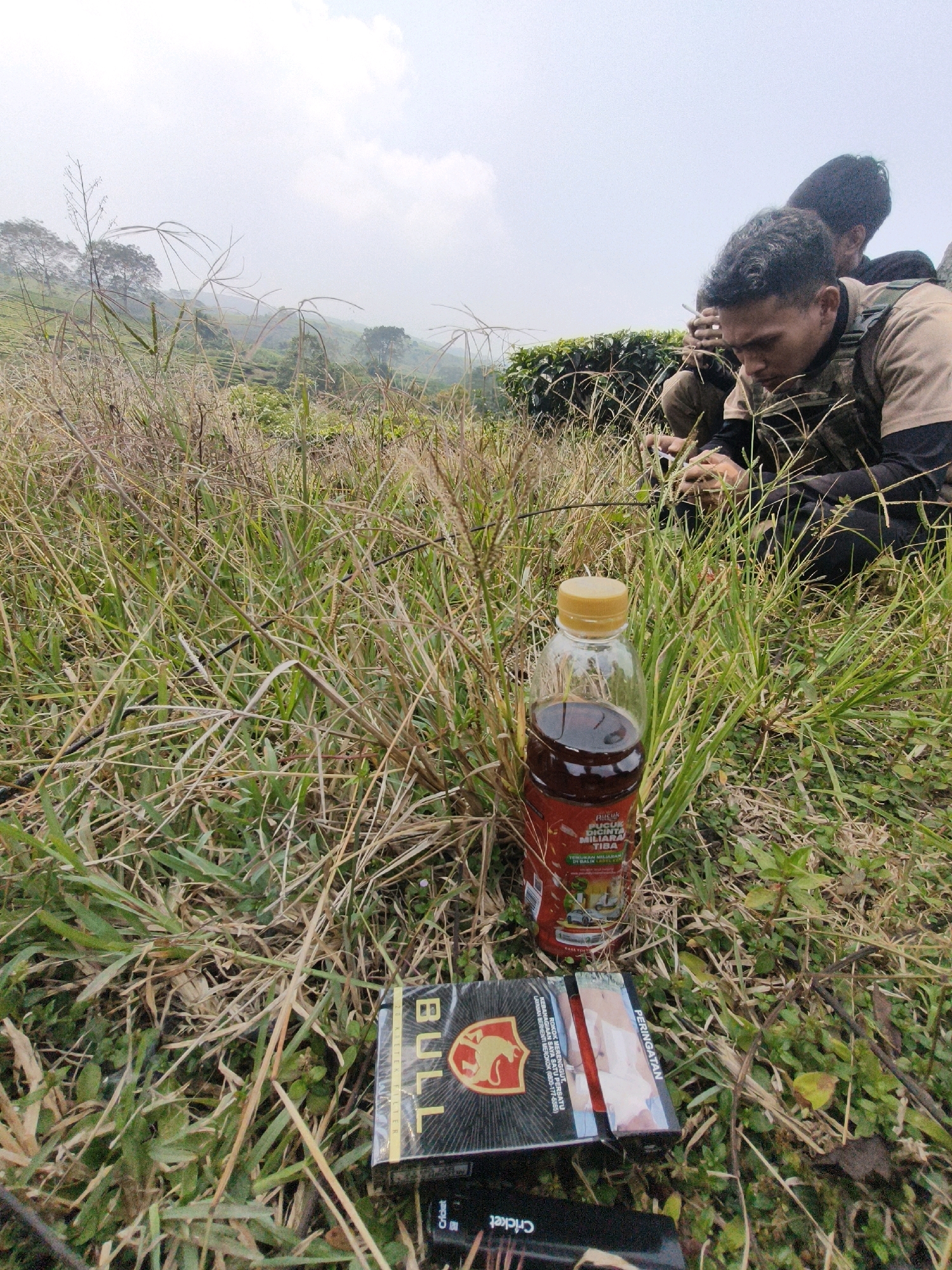 In an outdoor setting with dense foliage and a hazy sky, two individuals are present. One person, in the foreground and to the right, is wearing a camouflage vest over a brown long-sleeved shirt and appears to be focused on something in their hands. A second person is partially visible behind them, also looking down. In front of the person in the foreground, there is a bottle of dark liquid with a yellow cap and a pack of "BULL" cigarettes. A lighter is also visible on the ground next to the cigarette pack. The background shows a sloped landscape with green vegetation, possibly tea plantations, and trees in the distance. The overall impression is of a moment of rest or activity amidst a natural, somewhat overgrown environment, likely in a rural or semi-rural area. It is not possible to determine the exact location within the city of Pasar Minggu, Indonesia, from the visual information provided.
