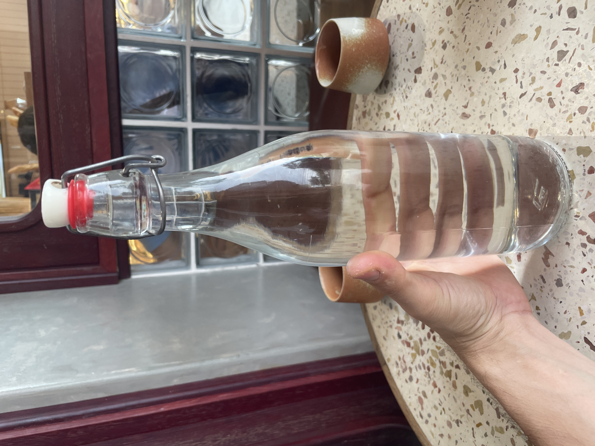 A close-up, rotated image captures a person's right hand holding a clear glass bottle filled with a transparent liquid, likely water. The bottle features a swing-top closure with a white stopper, a red rubber gasket, and a silver metal clasp. Faint embossed text, possibly "LG" or a logo, is visible near the base of the bottle.
The bottle and hand are positioned over a light-colored, speckled table surface, resembling terrazzo. To the upper right, a small, cylindrical ceramic cup with a mottled light brown and white finish rests on the table.
In the background, a dark wooden frame or panel runs vertically, partially obscuring a wall or partition made of square glass blocks. These blocks exhibit varying levels of transparency and reflections. Below this, a light-toned surface, possibly a window sill, is visible.
The scene appears to be an indoor or semi-enclosed setting, such as a cafe or restaurant, illuminated by natural light during the daytime. The location is Bac Nha Trang Ward, Vietnam. No specific activity beyond holding the bottle is evident.