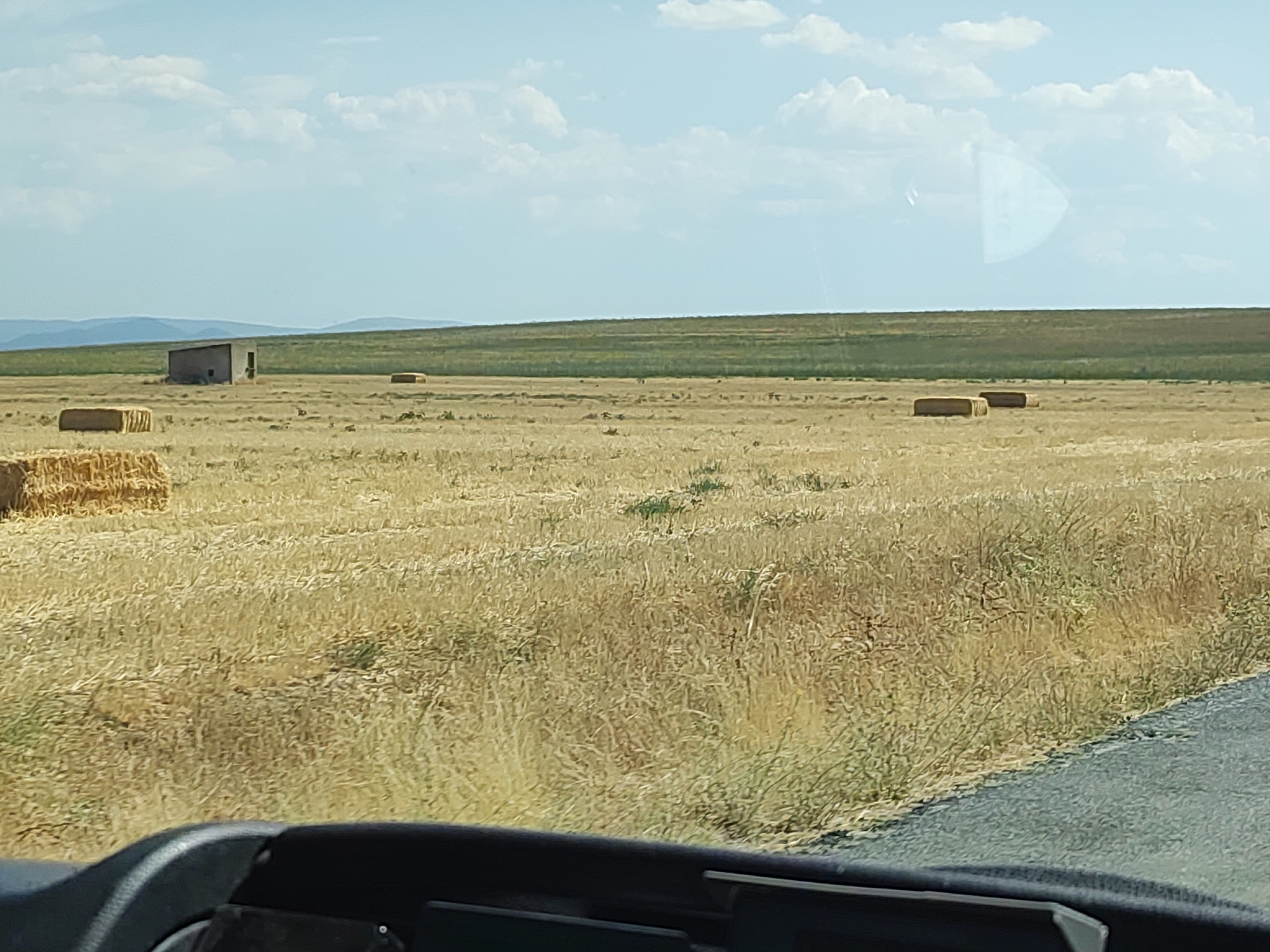 The image depicts a rural landscape under a sky with scattered clouds. In the foreground, the dashboard of a vehicle is visible, suggesting the photo was taken from inside. Beyond the dashboard, a dry, grassy field stretches into the distance, dotted with several rectangular bales of hay. To the left, a stack of hay bales is partially visible. In the middle ground, a small, weathered, rectangular structure with a slanted roof and an open doorway stands in the field. Further in the distance, rolling green hills form the horizon. The scene appears to be a cultivated field, likely harvested, given the presence of hay bales. The environment is dry and arid. The location is identified as Santa María la Real de Nieva, Spain.