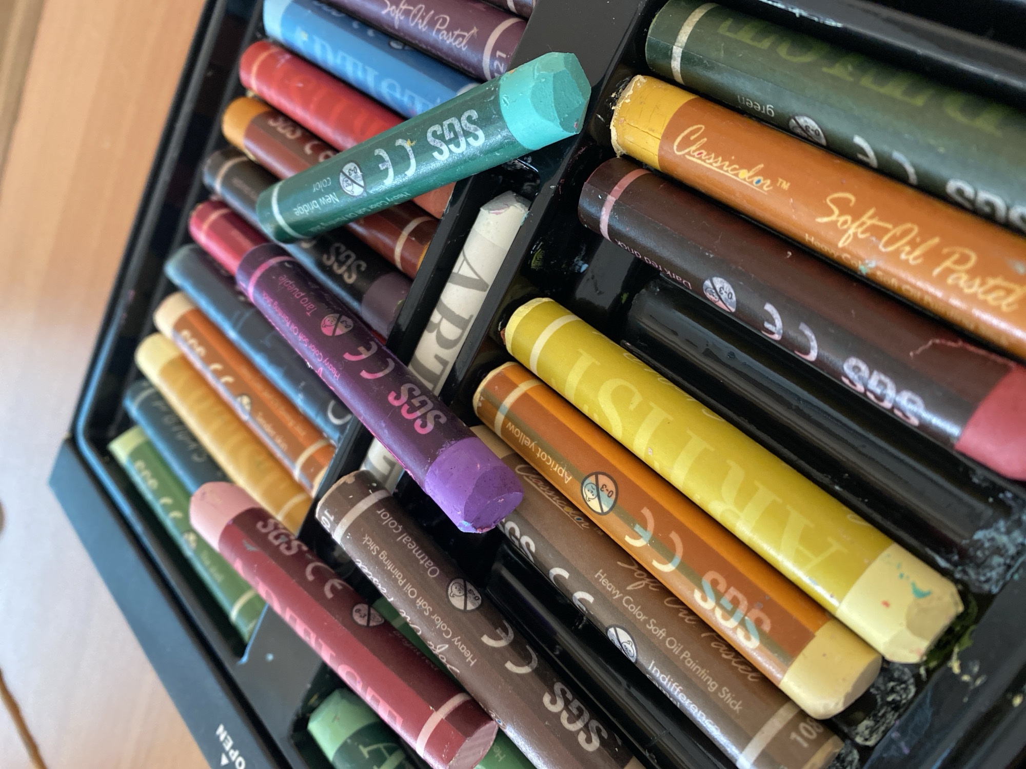The image shows a close-up view of an open box of oil pastels. The pastels are arranged in rows within the box's compartments. The pastels have a variety of colors including shades of brown, green, red, blue, yellow, purple, and white. Some of the pastels are labeled with text, including brand names like "Classicdor" and the words "Soft Oil Pastel", "Heavy Color Soft Oil Painting Stick," and color names like "Oatmeal color," "Apricot yellow," "Taro purple," "Tender skin," "New bridge," "Dark red drick," and "green." The "SGS CE" mark is visible on many of the pastels. The box appears to be made of black plastic. The image provides no information to infer a location in Yekaterinburg, Russia.