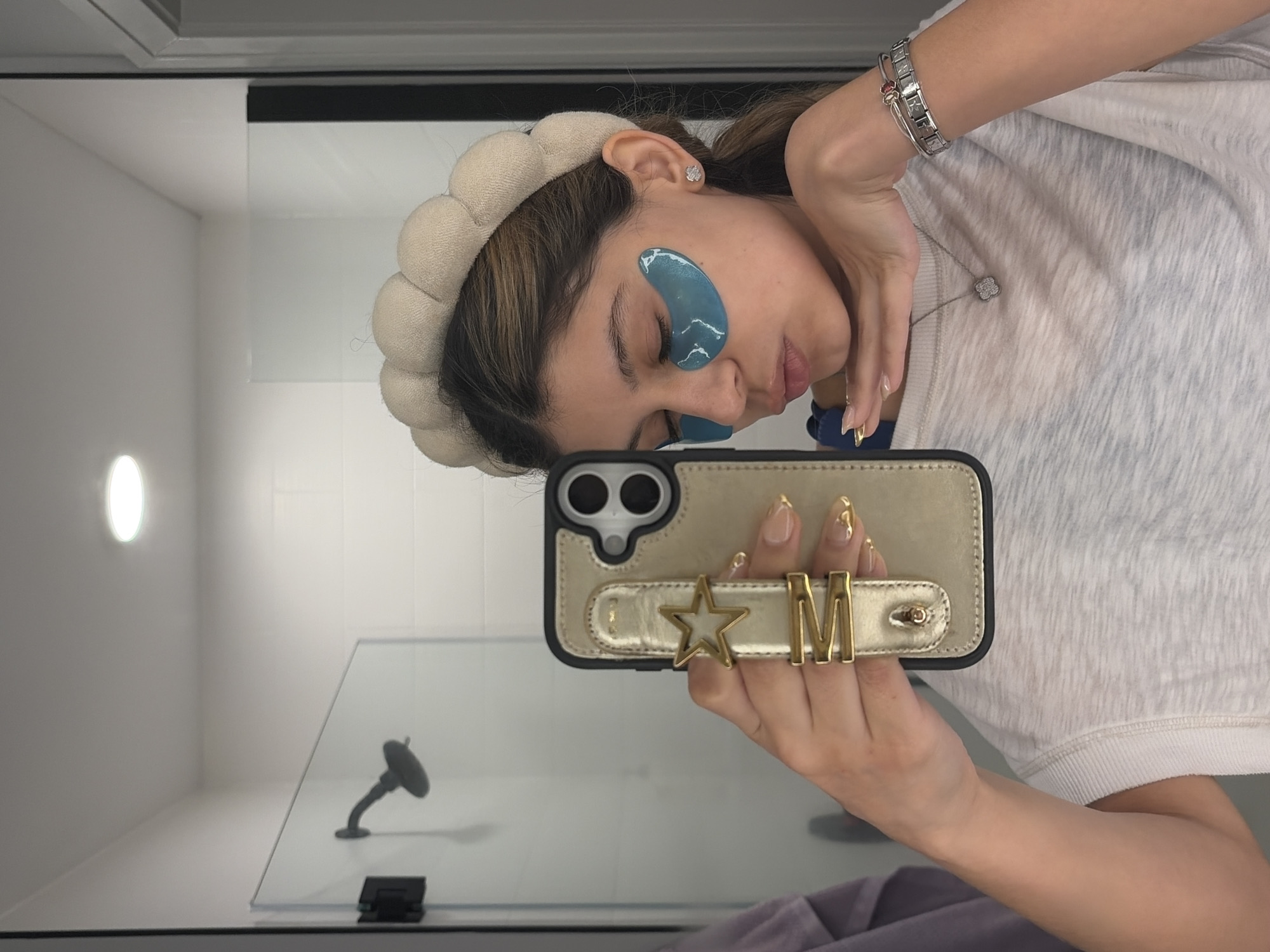 A woman in Austin, United States, is taking a selfie in what appears to be a bathroom. She has fair skin and wears a beige padded headband and blue under-eye patches. She holds a gold-colored iPhone with a black border, which features a gold strap adorned with a star and the letter "M". Her nails are long and have a gold design. The woman wears a silver bracelet on her wrist and a silver necklace with a clover-shaped pendant. She is dressed in a light-colored shirt. The background reveals a shower with a glass door and a minimalist shower head. A small, round light fixture is visible on the white wall. The overall environment is clean and well-lit.