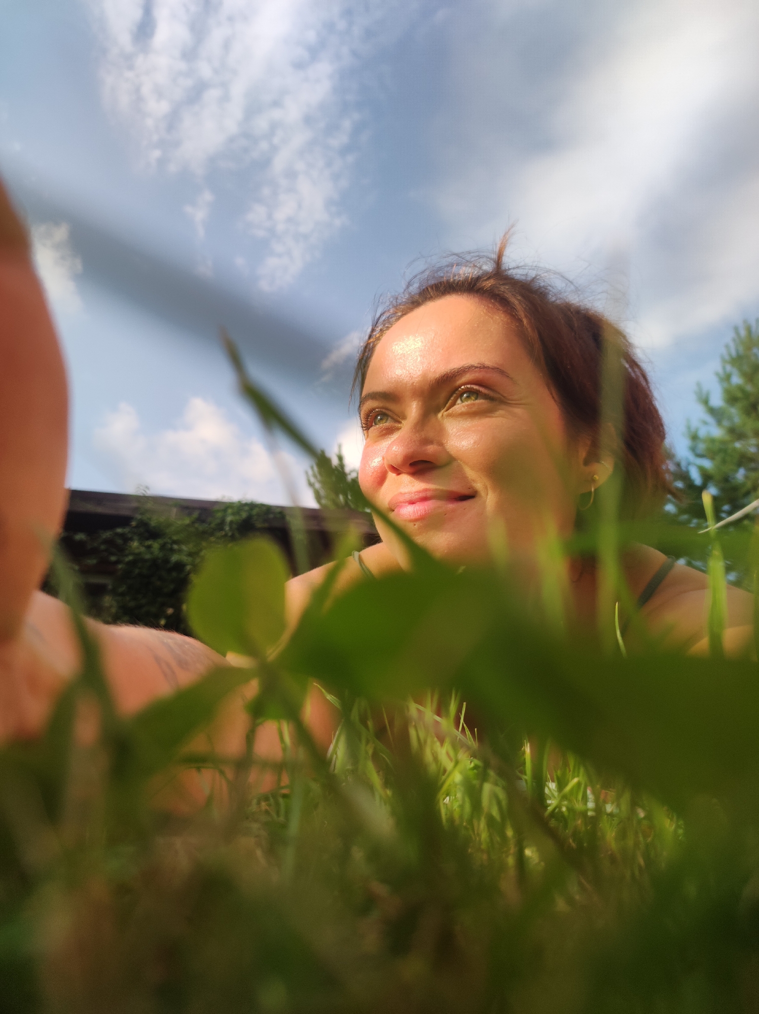 The image is a low-angle shot taken outdoors, likely in a grassy area, with a focus on a young woman looking up and to the right. Her face is well-lit by sunlight, highlighting her features and creating a natural glow on her skin. She has brown hair, parted and slightly messy, and green eyes. She is smiling faintly, with a relaxed and content expression. She appears to be wearing a dark-colored top.
In the foreground, large blades of green grass are blurred, creating a natural frame and emphasizing the perspective. In the background, a fence or wall is visible, covered in green foliage, suggesting a garden or yard. Beyond that, there are trees and a bright blue sky with scattered white clouds. The overall impression is one of a sunny day, possibly in a suburban or rural setting. The woman's left hand and arm are partially visible in the lower left corner, also slightly out of focus.
There is no information available to suggest this image was taken in the city of Фоки, Russia.