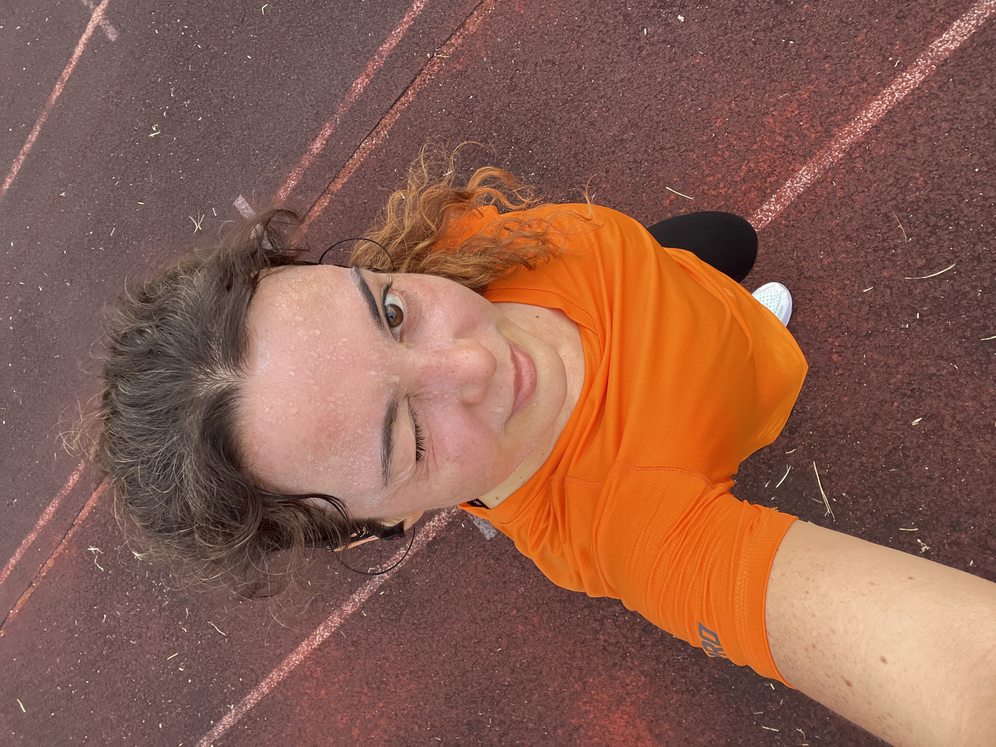 A woman with curly brown hair, wearing an orange t-shirt and black leggings, is winking at the camera while standing on a red running track. She has sweat on her forehead and cheeks, suggesting she has been exercising. The track has white lines marking lanes. The image appears to be a selfie taken from a high angle. The background suggests an outdoor athletic facility, possibly in the city of Phan Thiết, Vietnam, given the context.