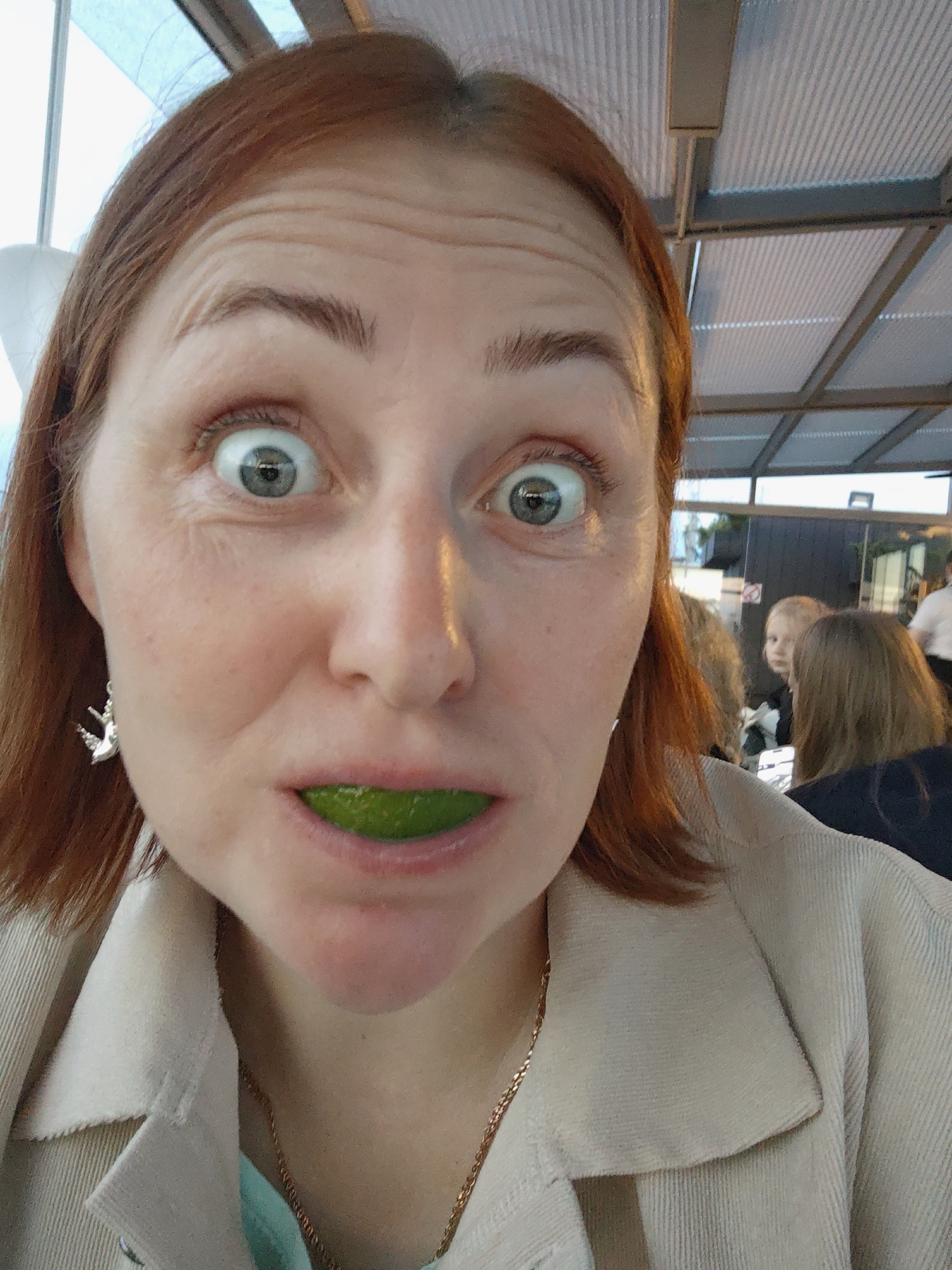 A woman with auburn hair is taking a selfie, holding a slice of lime in her mouth. Her eyes are wide open and she is looking directly at the camera. She is wearing a beige jacket over a teal shirt and a gold chain necklace. A silver earring with a charm is visible on her left ear. In the background, other people are seated at tables under a corrugated roof structure, suggesting an outdoor seating area of a restaurant or cafe. The lighting indicates it is daytime. The scene is likely in Saint Petersburg, Russia, given the request.