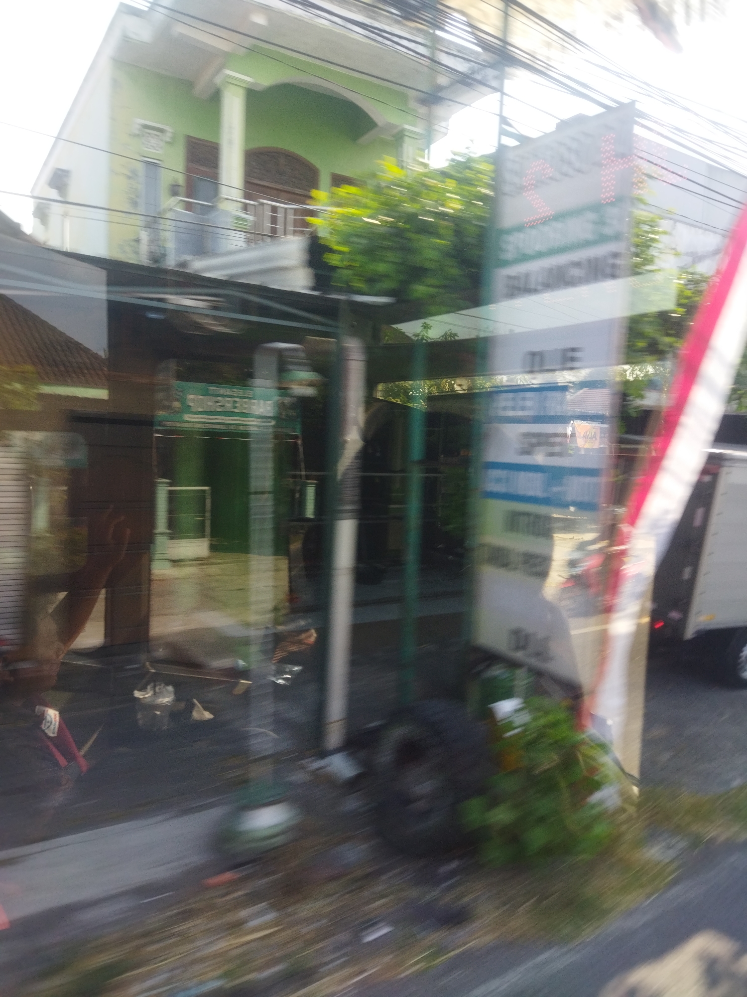 The image depicts a street view in the city of Begajah, Indonesia, with a focus on the facade of a building and reflections on a glass surface. The building, painted in light green and white, features a covered porch with white pillars and ornate wooden doors. Lush green trees are visible in the background. The foreground is dominated by reflections on a glass pane, showing a red and white striped object, a white truck, and various signs. One sign, with red illuminated text, appears to advertise a business, with visible words like "STOKING" and "BALANCING". Another sign, partially obscured, has blue and white lettering. The ground is paved and shows signs of wear and some scattered debris. The overall impression is that of a busy street scene, possibly viewed from inside a vehicle.