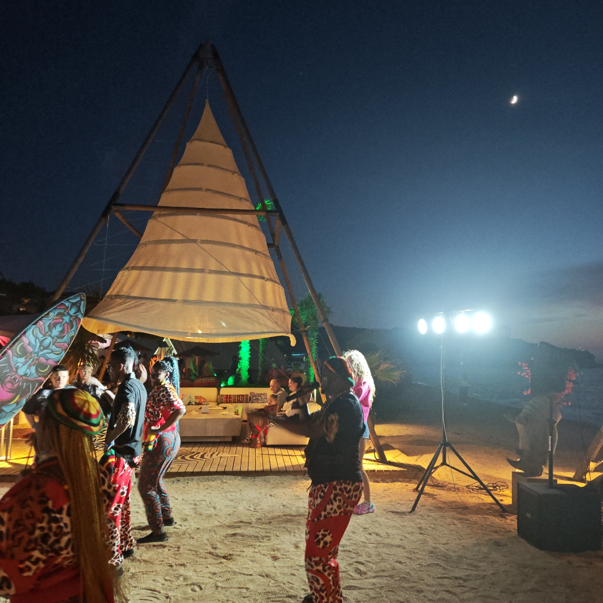 In Мысовское сельское поселение, Ukraine, at night, a group of people are gathered on a sandy beach. In the background, a large, triangular white tent structure is supported by wooden beams. Several individuals are dancing, some wearing vibrant, patterned clothing, including animal prints and geometric designs. One person is playing a drum. Other attendees are seated at tables or standing around the tent. Green lights illuminate some of the foliage behind the tent. A surfboard with a colorful design is leaning against a pole on the left side of the image. In the distance, a crescent moon is visible in the dark blue sky, and bright spotlights illuminate the scene. The overall atmosphere appears to be a lively outdoor event.