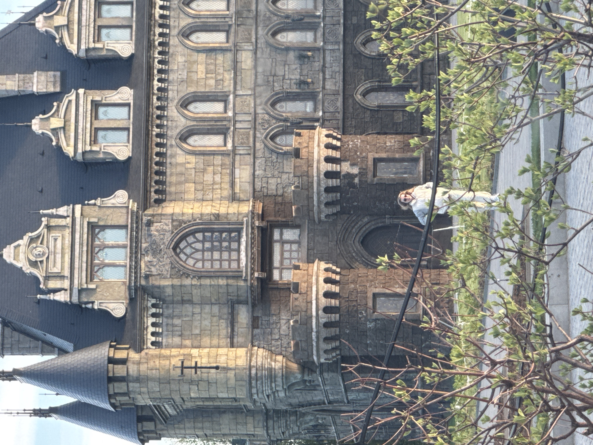 The image shows a section of a medieval-style castle building with turrets, windows, and stone walls. A person stands in front of the building. The location appears to be in a city, but its name is unspecified.