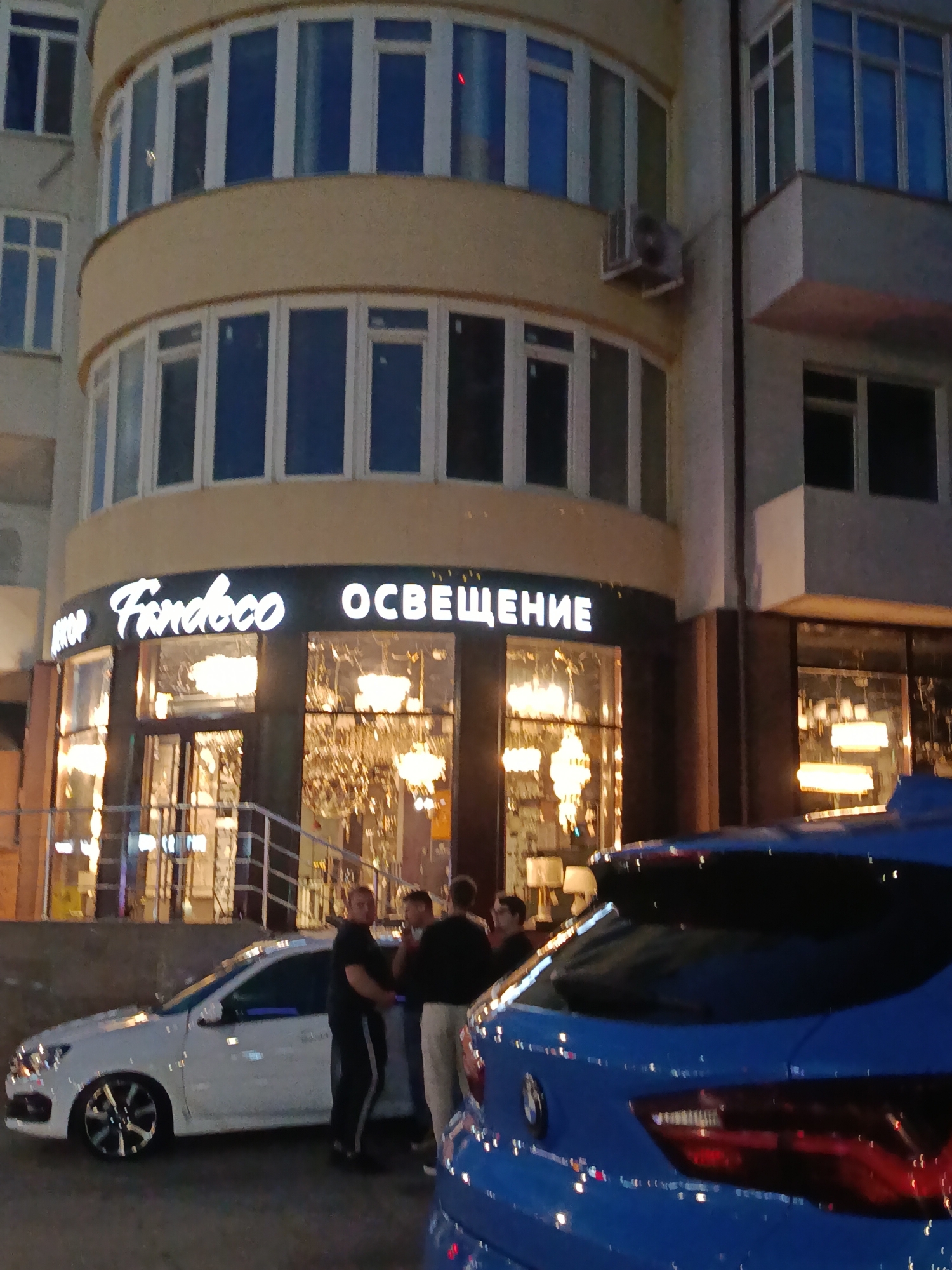 The image depicts the exterior of a building at night, likely a store. A prominent sign above the entrance reads "Fardeco ОСВЕЩЕНИЕ" (Fardeco Lighting), indicating the establishment sells lighting fixtures. The store windows display several chandeliers and other decorative lights, suggesting it is a lighting showroom.
In front of the store, a group of men are gathered, seemingly engaged in conversation. One man in a black t-shirt and dark pants stands near the entrance, while others are positioned slightly further away. A white car is parked to the left of the entrance, and the rear of a blue car is visible in the foreground to the right.
The building features multiple floors with numerous windows. The overall setting appears to be a street in a city, with the warm glow of artificial lights illuminating the scene. The image does not provide explicit information about the city of Saratov, Russia, but it is consistent with an urban environment at night.