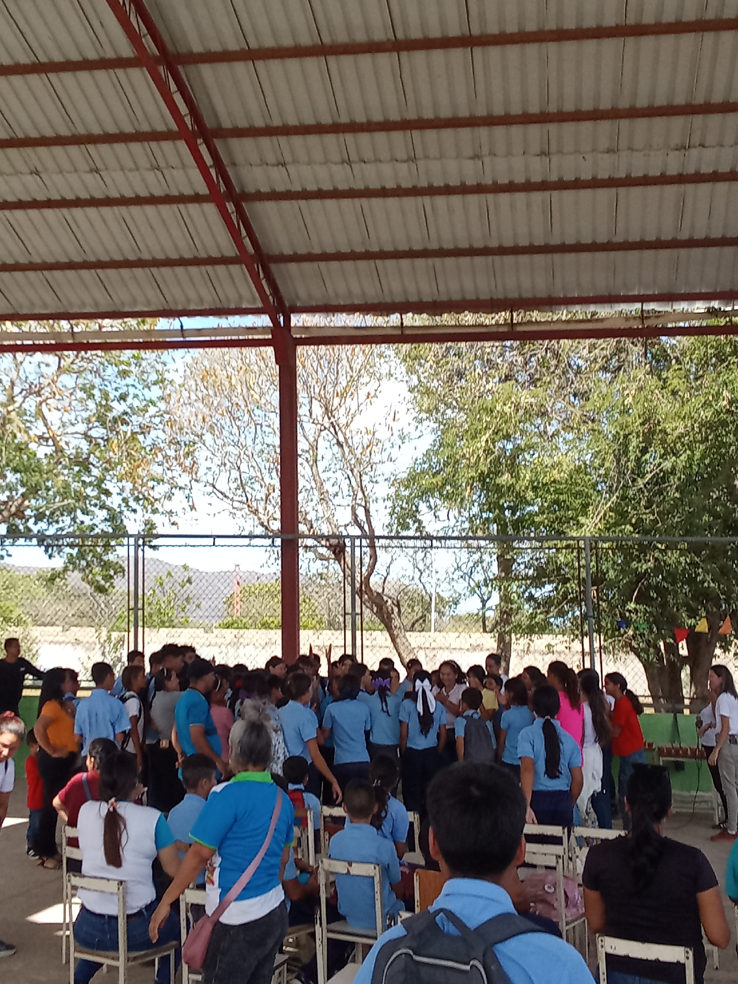Under a corrugated metal roof supported by a red steel frame, a large group of people, primarily children wearing blue shirts, are gathered. The children stand in the center of the space while adults are seated on white chairs or stand along the edges. Some children raise their hands. A chain link fence is visible in the background, with trees and a distant mountain range beyond. The location is Juan Griego, Venezuela.