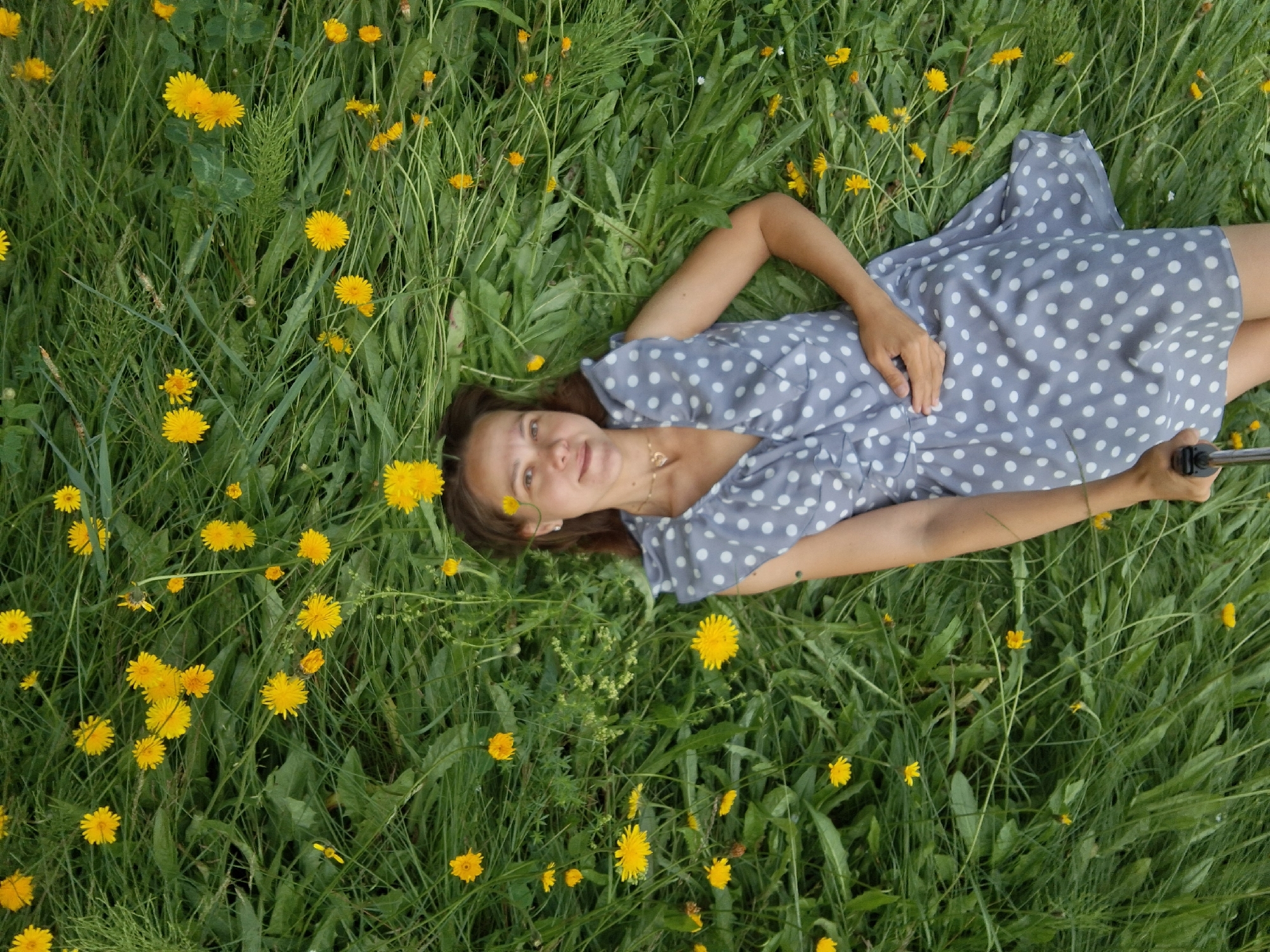 A young woman with brown hair lies on her back in a field of green grass and yellow dandelions. She is wearing a light blue dress with white polka dots. Her head is tilted back, and she is looking up at the camera with a slight smile. Her right arm is bent at the elbow, and her hand rests on her stomach. A black selfie stick is visible in the lower right corner of the image, held by her right hand. The background is filled with lush green grass and scattered yellow dandelion flowers, creating a natural and serene outdoor setting. It is not possible to determine the location within the city of Chaikovsky, Russia from the image alone.