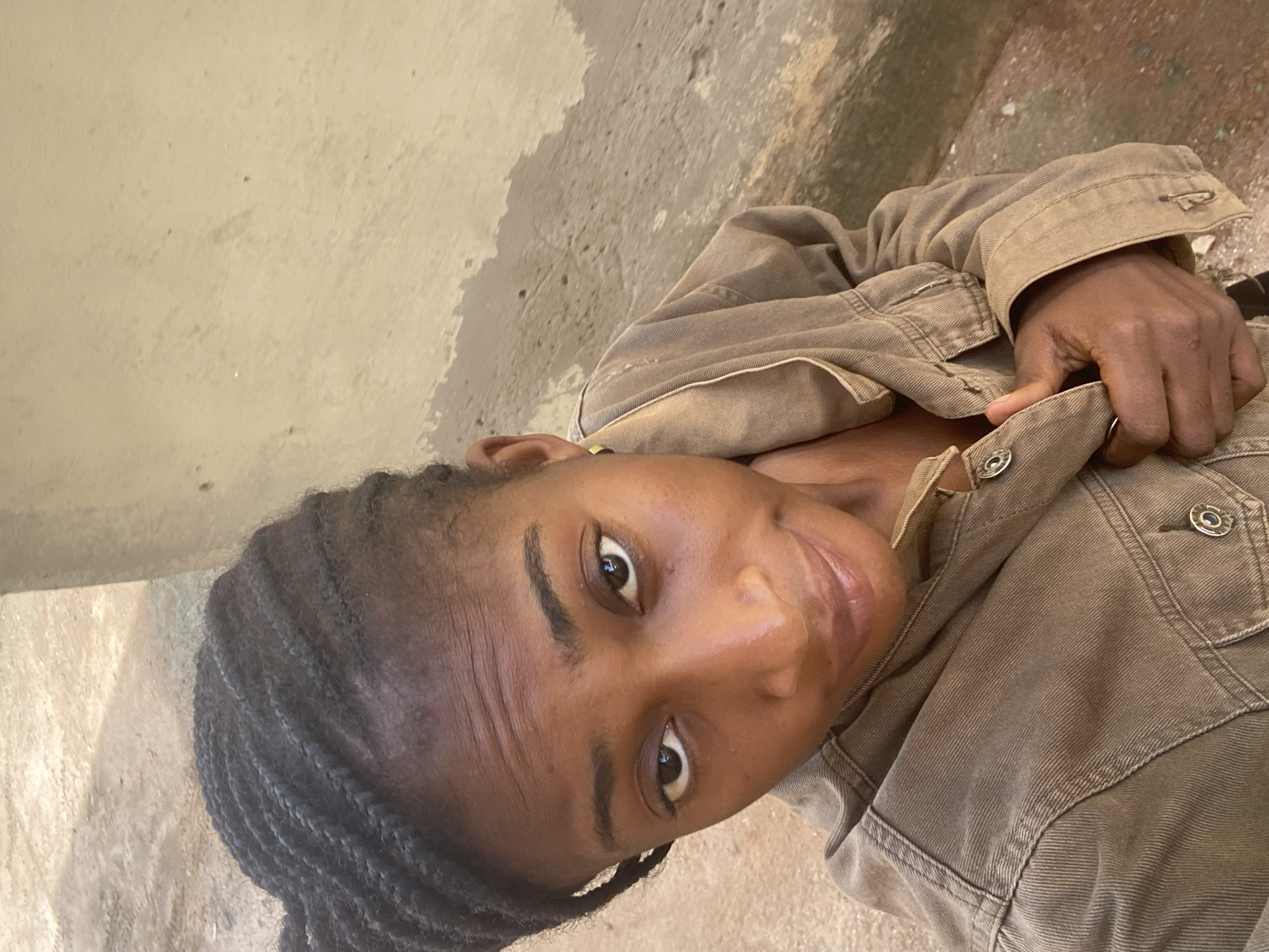 A female individual with a dark complexion and black braided hair is centered in an outdoor, upward-angle shot within an urban setting in Abuja, Nigeria. She wears a light brown or khaki-colored denim jacket with visible stitching and silver snap buttons, holding the jacket's front lapel with her right hand. Her eyes are dark and look directly towards the camera, with a slight, subtle smile on her lips. Faint horizontal lines are visible across her forehead. A small, golden stud earring is present in her left ear. The background consists of a light beige or tan wall on the right, displaying areas of darker discoloration consistent with dampness or age, and a dusty, light brown, unpaved ground surface on the left and foreground. Natural lighting illuminates the scene uniformly.