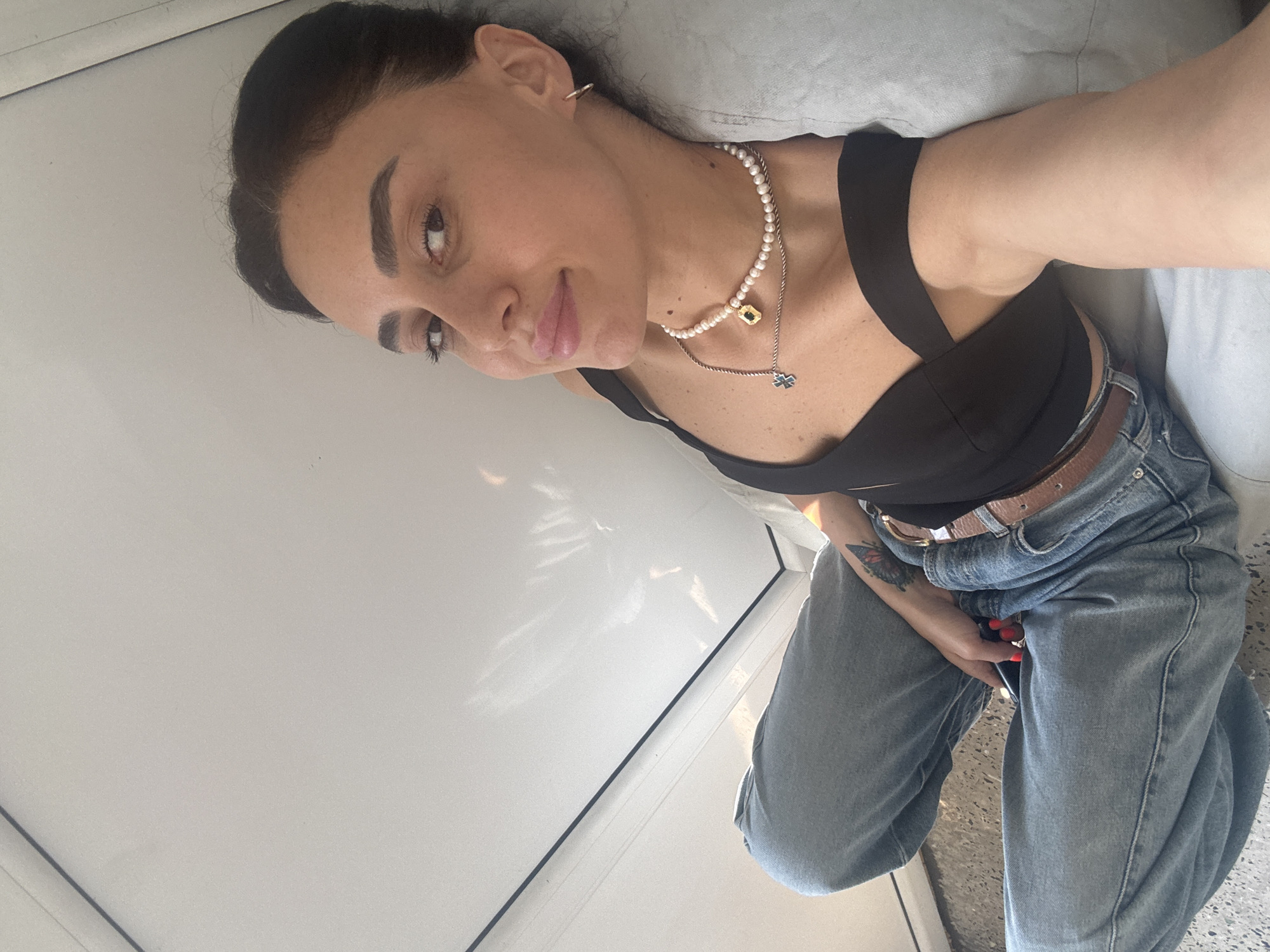 A woman with dark hair, wearing a black crop top, blue jeans, and a pearl necklace, sits with her legs crossed. She is leaning against a light blue pillow. Her right arm is extended, and she holds a phone in her hand. A tattoo of a butterfly is visible on her left arm. The background appears to be an indoor setting with white walls and a light gray floor. The lighting suggests it is daytime, with sunlight casting shadows on the wall. The specific location in Tbilisi, Georgia, is not identifiable from the visual information.