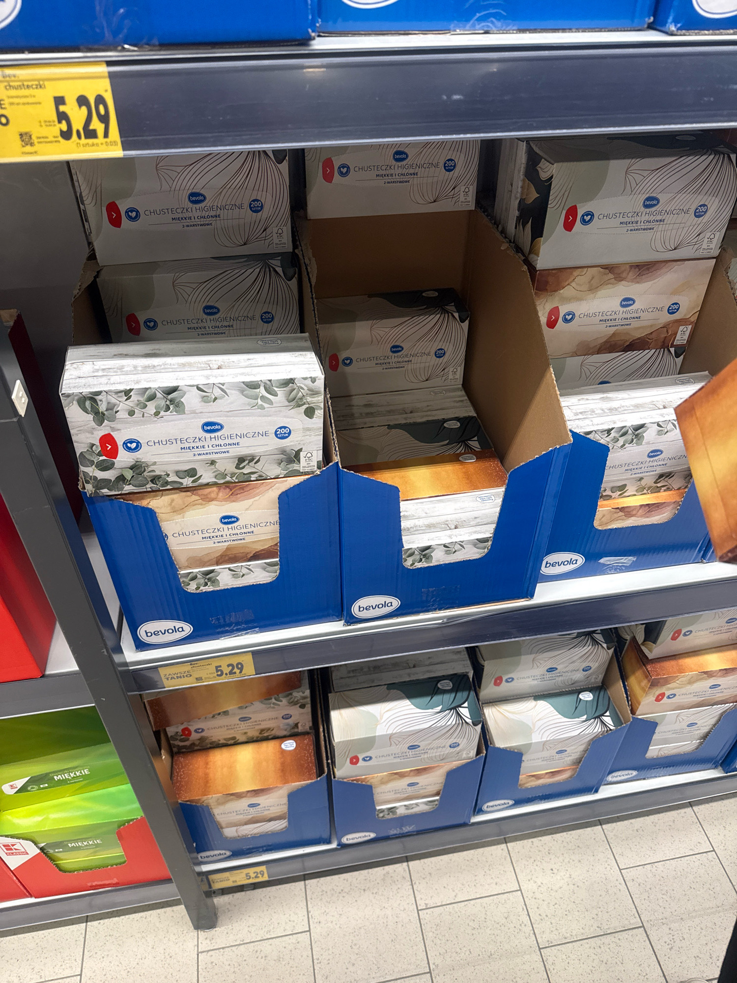 The image shows a retail shelf display of boxed facial tissues. The tissues are branded "bevolá" and are described as "CHUSTECZKI HIGIENICZNE MIĘKKIE I CHŁONNE 2-WARSTWOWE" (hygienic tissues, soft and absorbent, 2-ply). The boxes feature a stylized line drawing design of what appears to be fan-like shapes or abstract floral elements in white and gray, with some boxes also showing a green eucalyptus leaf design and others a gradient of brown and gold. The tissues are packaged in cardboard boxes stacked within larger blue cardboard display units.
A yellow price tag with the number "5.29" is visible in the lower portion of the image, indicating the price of the tissues. The overall environment suggests a supermarket or discount store aisle. The tiled floor visible at the bottom of the image is consistent with retail flooring.
Although the image is of a retail display, without additional context or explicit visual cues within the image itself, it is not possible to determine the specific city of Gorzów, Poland, as the location. The image focuses solely on the product display.