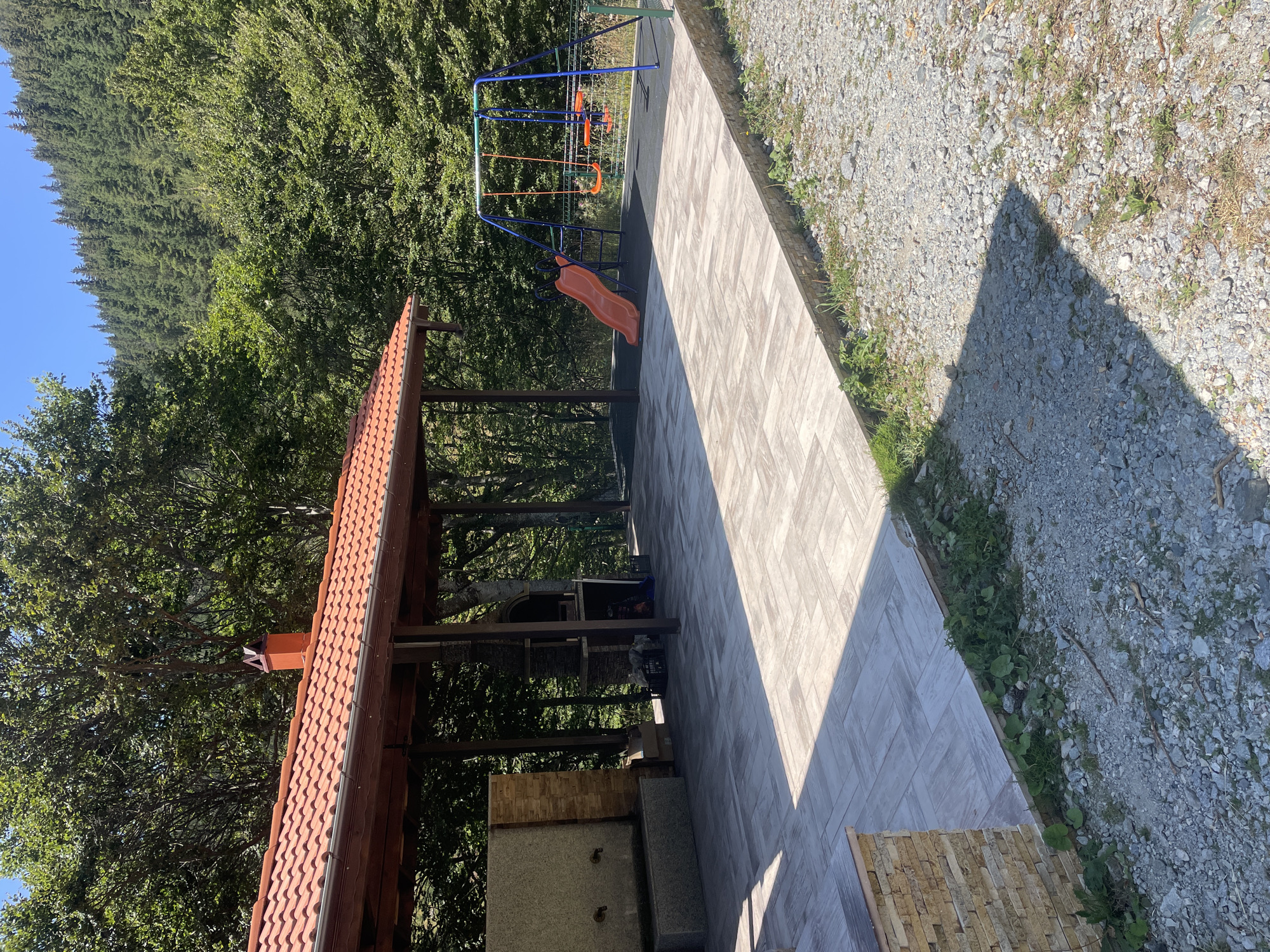 The image depicts an outdoor recreational area nestled within a forested environment, likely in or around Bosilkovo, Bulgaria. The scene is dominated by a covered pavilion with a reddish-brown tiled roof and exposed wooden beams. Beneath the pavilion, a stone fountain with two spigots is visible, adjacent to a brick structure. Further inside, a barbecue area is set up, with a stone chimney and grill.
The ground in front of the pavilion is paved with large, light-colored rectangular tiles arranged in a herringbone pattern, extending outwards and connecting to a gravel path. To the right of the tiled area, a children's playground is situated, featuring a blue swing set with two orange seats and an orange slide. The playground area is set on a dark rubber matting.
The background is characterized by dense, lush green trees forming a forest that extends up a hillside, with a clear blue sky overhead. The overall impression is that of a tranquil, natural setting prepared for outdoor gatherings and family activities. There are no people visible in the image.
