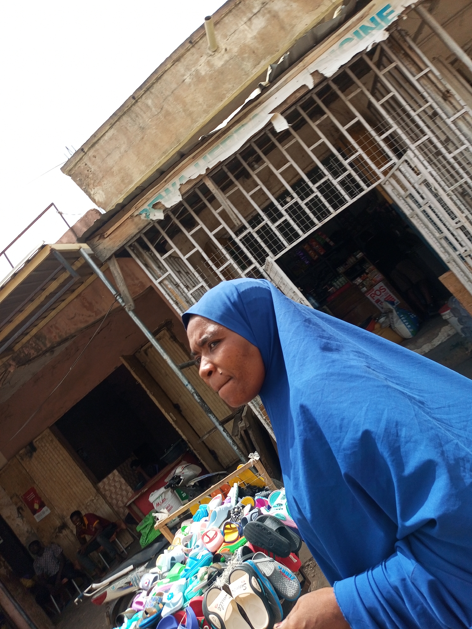 A woman wearing a blue hijab is standing in front of a pile of footwear for sale in what appears to be a market area. The setting seems to be Arawa, Nigeria, based on the general visual cues of the environment. Behind her are commercial buildings, with the top portion of one having a 'CINE' sign visible. A second building has an open entrance area with two men sitting inside. The pile of footwear in the foreground includes various colors and styles of sandals. The overall lighting suggests it is daytime.