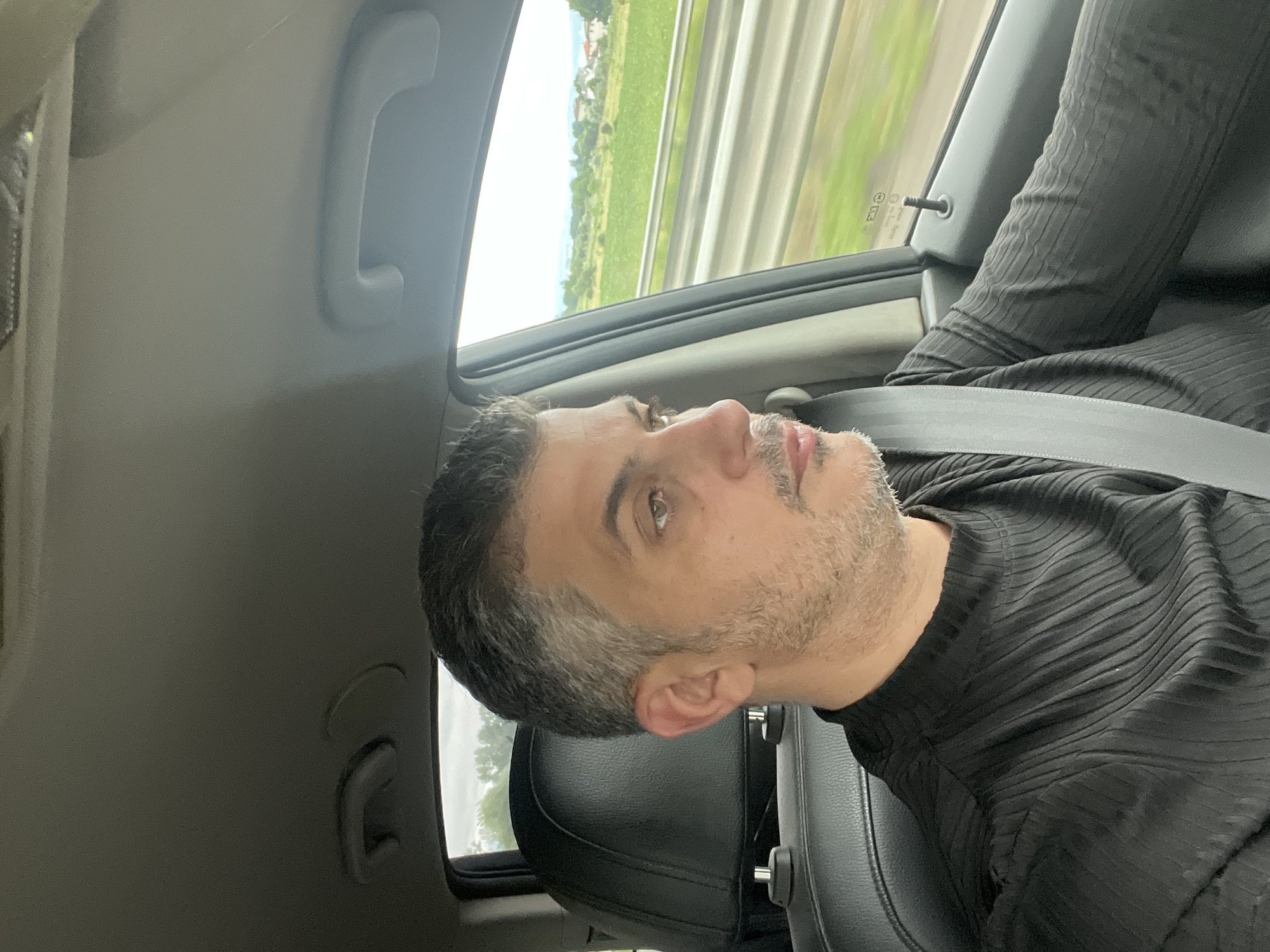 A middle-aged man with short, salt-and-pepper hair and a light beard is driving a car. He is wearing a black, ribbed, mock-neck shirt and a grey seatbelt. The interior of the car is visible, including the black leather seat and the overhead handle. Through the passenger window, a blurred green landscape with some houses can be seen, indicating the car is in motion. The lighting suggests it is daytime. The location is Unknown, Unknown.
