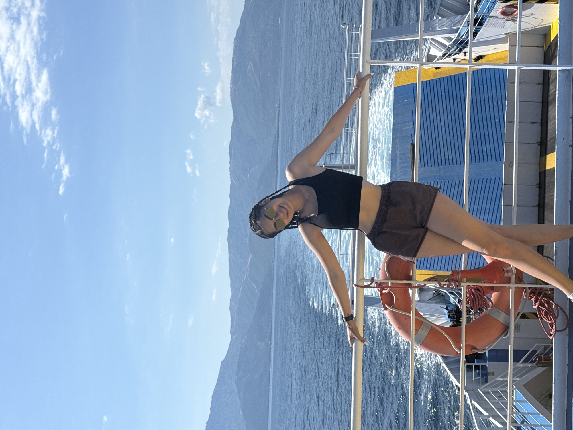 A young woman with braided hair and sunglasses smiles as she stands on the deck of a ferry, leaning against a white railing. She wears a black crop top and brown shorts. The ferry is moving through blue water, with a wake trailing behind it. In the background, a mountainous coastline under a clear blue sky is visible. A life preserver hangs on the railing to her left. The scene appears to be in Greece, given the mountainous coastline and the presence of a ferry.