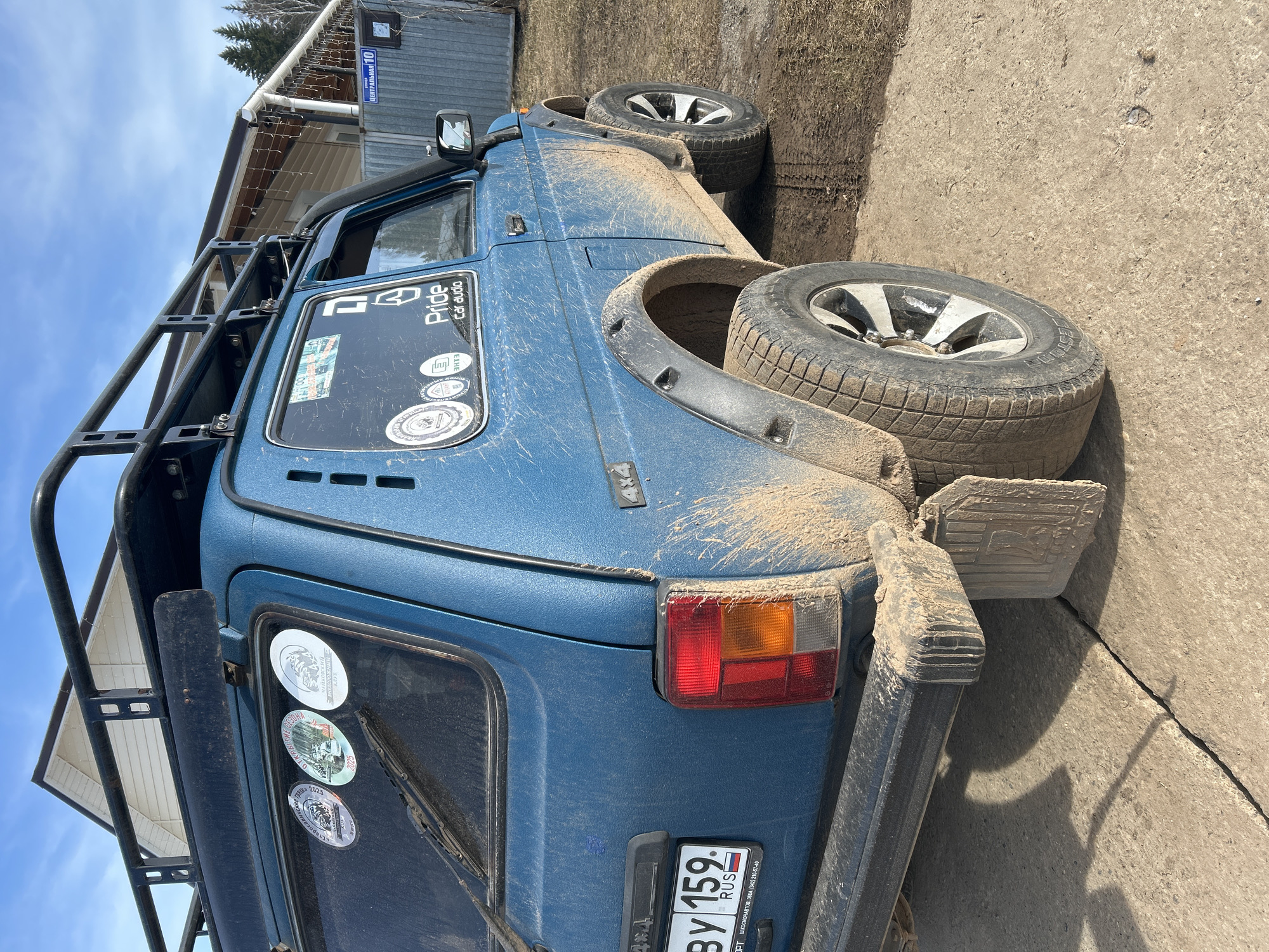 The image shows a blue Lada Niva SUV, heavily covered in mud, parked on what appears to be a concrete or paved area. The vehicle has a roof rack and spare tire mounted on the rear. Several stickers are visible on the rear windows, including ones related to off-roading events in 2025, and "Pride Car Audio" on the side window. The license plate reads "By 159" and indicates the vehicle is registered in Russia. Street sign visible reads "ЦЕНТРАЛЬНАЯ 10". The location is likely in the city of Уральское, Russia.