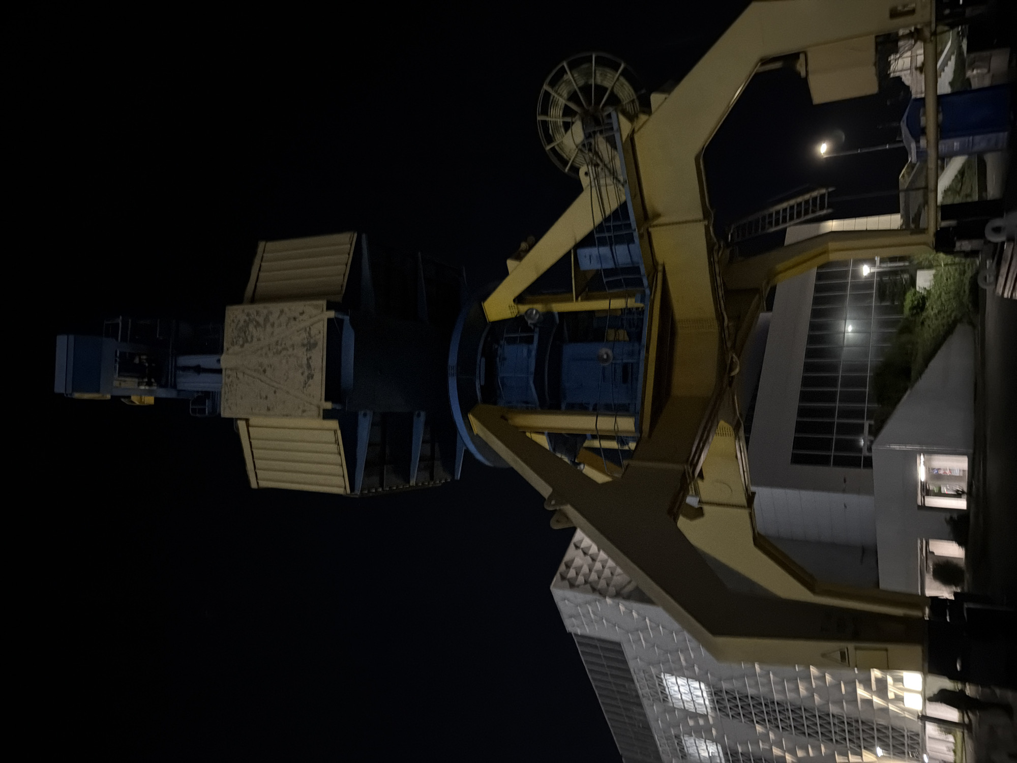 The image depicts a large crane structure at night in Burgas, Bulgaria. The crane is constructed of yellow steel. It has a rotating hoist and a ladder leading to the top. Several buildings are visible in the background. Some buildings are white with reflective glass windows, and one building is a darker color. There is a streetlight in the background illuminating part of one of the buildings. The image appears to be rotated.