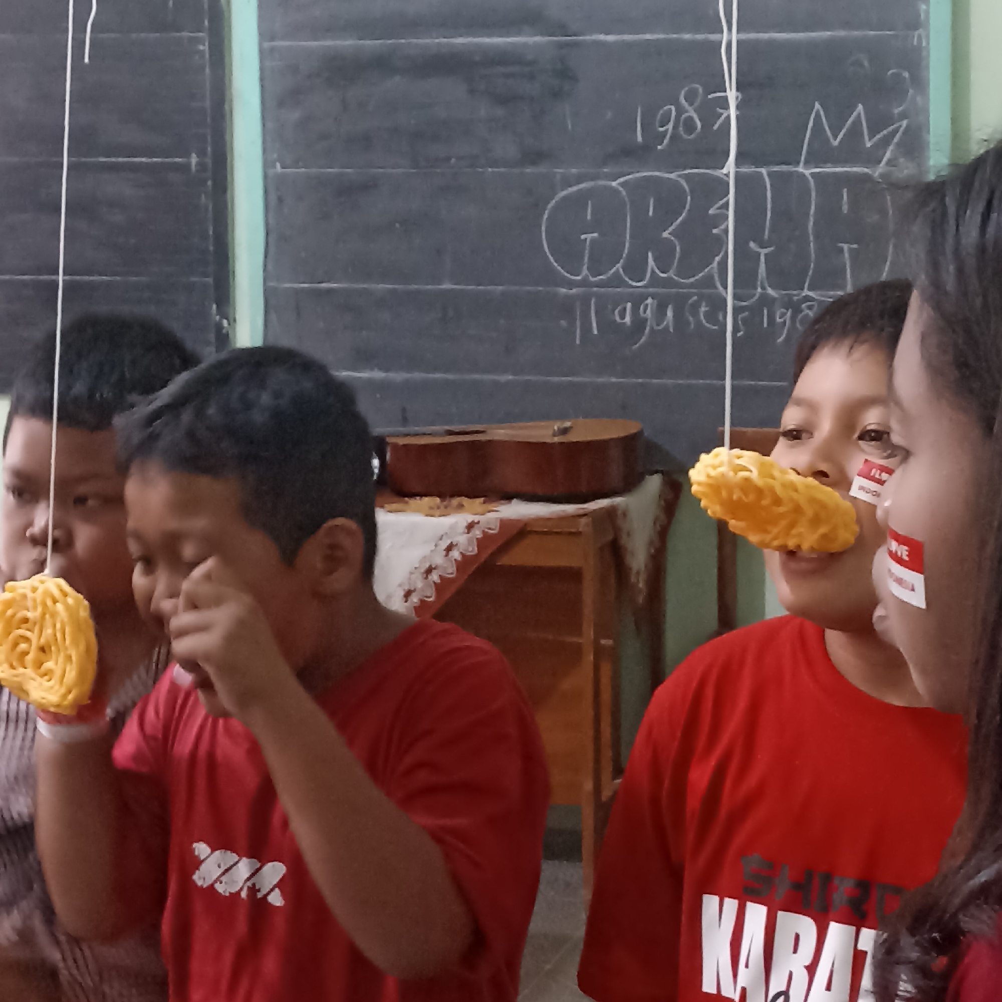 The image shows a group of children in red shirts participating in a game, possibly a competition, in what appears to be a classroom or community hall. The background features a dark chalkboard with white writing and drawings, including the numbers "1987", "AR", and "11 agustu s 198". There is also a crown-like drawing. A guitar is resting on a table covered with a patterned cloth.
In the foreground, three children are visible. Two boys are actively engaged in the game, holding yellow, circular snacks attached to strings, attempting to eat them without using their hands. The boy on the left is partially visible, and the boy in the middle is focused on the snack. Another child's face is visible on the right, with red and white stickers on their cheeks, resembling Indonesian flag colors, and also attempting to eat a similar snack. The children are wearing red t-shirts. The overall scene suggests a festive or celebratory event, likely an Independence Day celebration given the Indonesian flag colors and the date "11 agustu s 198" written on the chalkboard. The setting is likely within the city of Magelang, Indonesia.