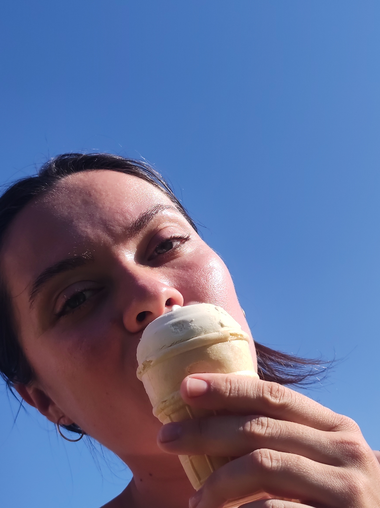 A person is seen from a low angle, against a clear blue sky. The individual, whose face is visible, is fair-skinned and appears to be eating an ice cream cone. The cone is held in their right hand, and the ice cream is white in color. The location is not evident in the picture.