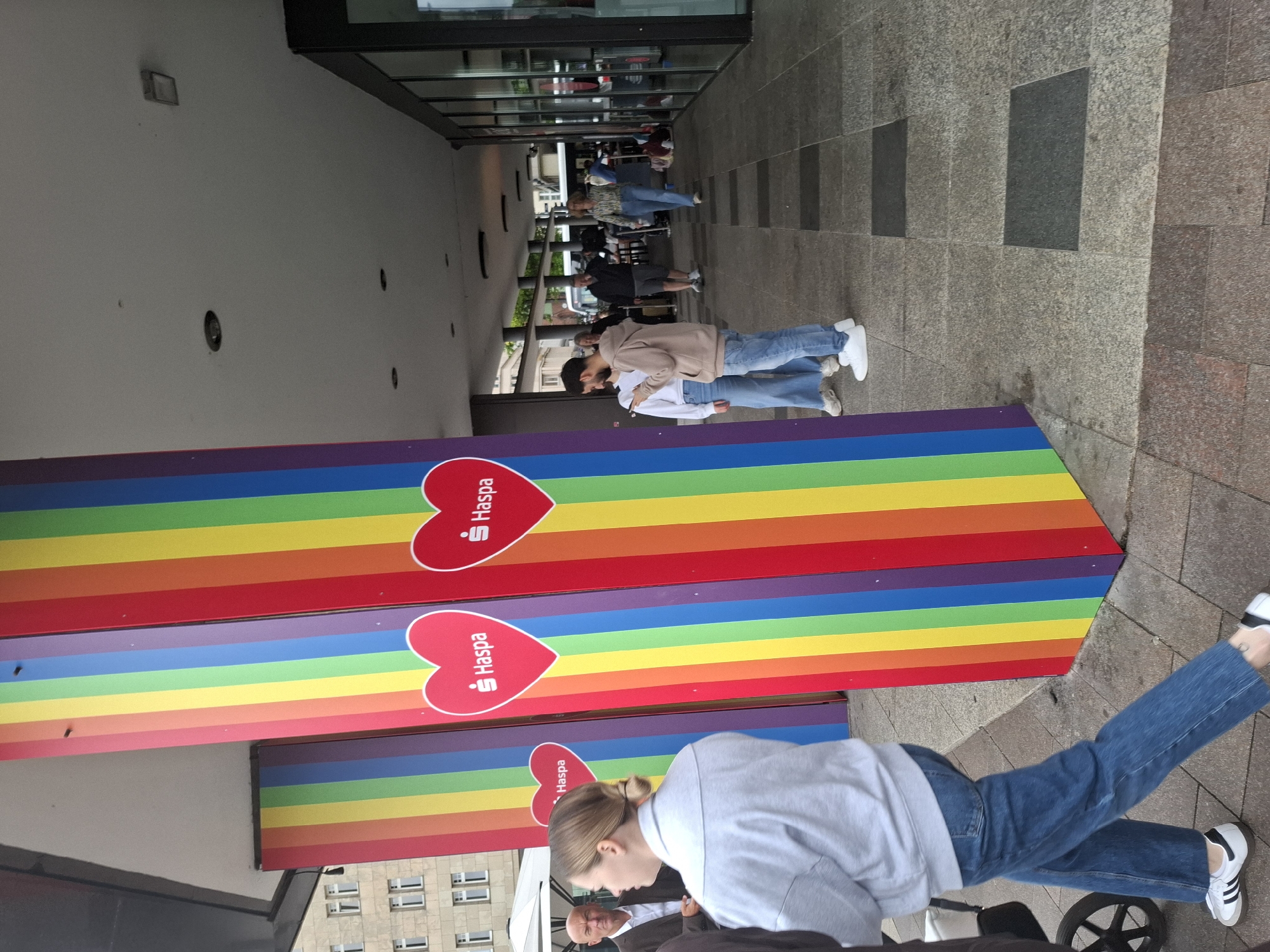 The image shows a street scene in Hamburg, Germany, where large, colorful rainbow flags are displayed on columns. Several people are present, including a woman in the foreground wearing a grey sweatshirt and blue jeans, and another woman in the background wearing a patterned shirt and blue jeans. A man in a suit is also visible. The setting appears to be an urban area with a building facade and a covered walkway. The rainbow flags, along with heart-shaped logos bearing the name "Haspa," suggest a Pride event or a corporate sponsorship related to LGBTQ+ awareness. The ground is paved with stone tiles.