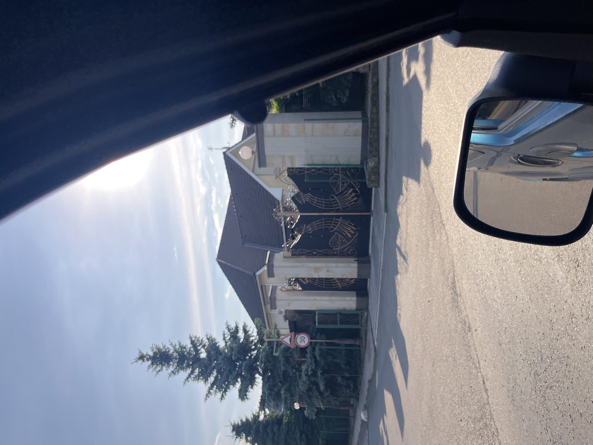 The image, taken from inside a vehicle, captures a residential street in Mynai, Ukraine, bathed in bright sunlight. In the foreground, a side-view mirror reflects a blue car. The middle ground features a paved road casting long shadows from an unseen object. On the right side of the road, a large, ornate black gate with golden decorative elements stands in front of a light-colored house with a dark roof. The gate is flanked by two tall, decorative pillars. To the left of the gate, a speed limit sign showing "30" is visible, along with a cautionary sign depicting a speed bump. Several tall evergreen trees line the property behind the gate. The sky is a clear blue with some wispy clouds and a bright sun creating a glare in the upper right corner of the image.