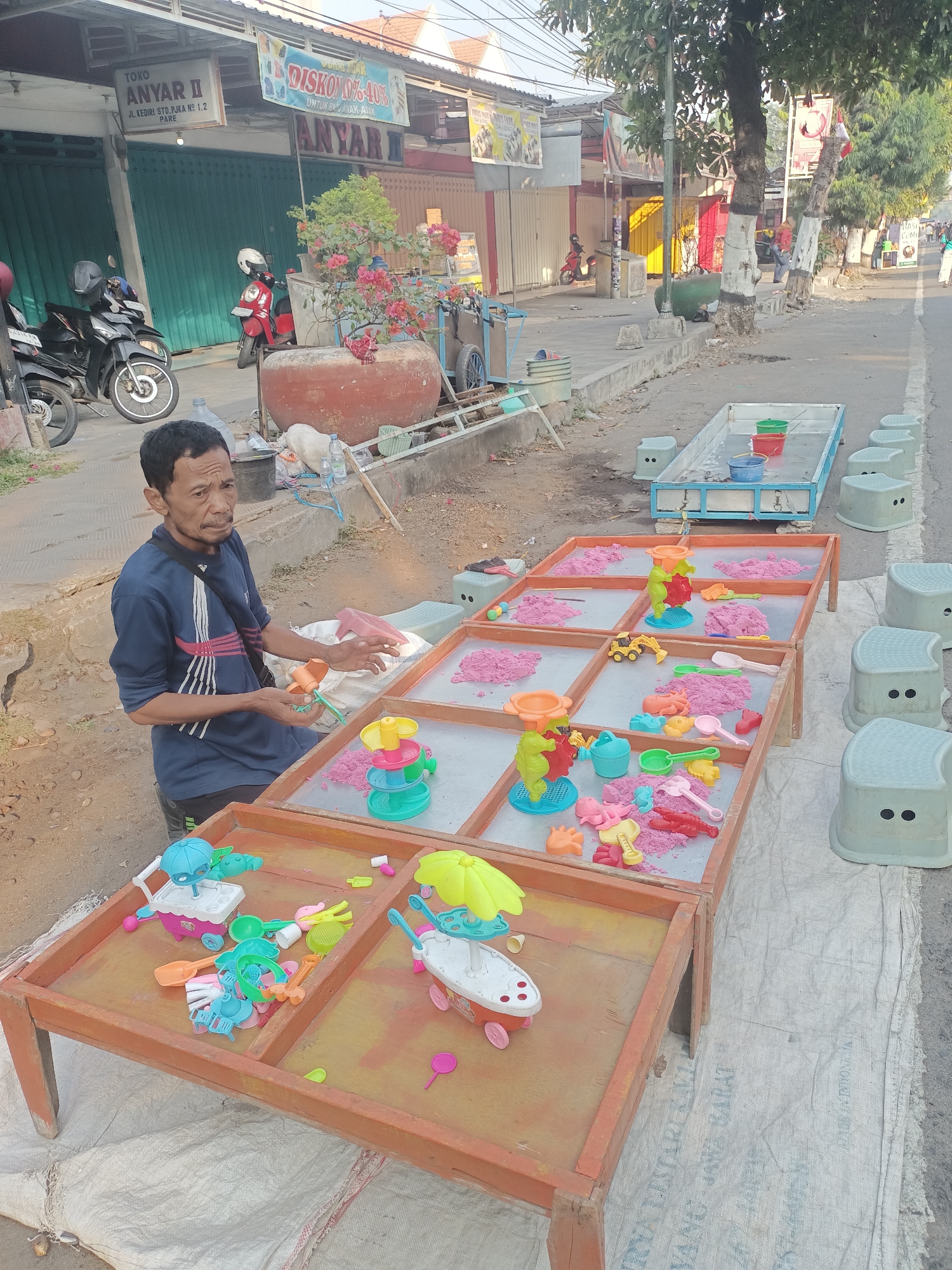 In Pare, Indonesia, a man sits in front of a display of colorful kinetic sand toys, likely engaged in their sale. The scene is set on a sidewalk next to a street, with a storefront visible in the background bearing the sign "TOKO ANYAR II JL. KEDIRI STD. PJKA NO 1.2 PARE". The storefront has green metal doors. In front of the store, motorcycles are parked. The man, wearing a blue shirt with red and white stripes and a black strap across his chest, holds a toy in his hands and gestures with his other hand, suggesting he is actively interacting with the toys or a potential customer.
The toys are arranged on wooden tables filled with pink kinetic sand. Various molds and plastic toys, including sea creatures, vehicles, and kitchenware, are scattered across the sand. Several light blue plastic stools are positioned around the tables, presumably for customers. A large water bottle and a white dog are also visible near the tables. In the background, more storefronts, trees, and street activity indicate a bustling urban environment. The overall impression is a typical street-side vendor setup in a commercial area of Pare.