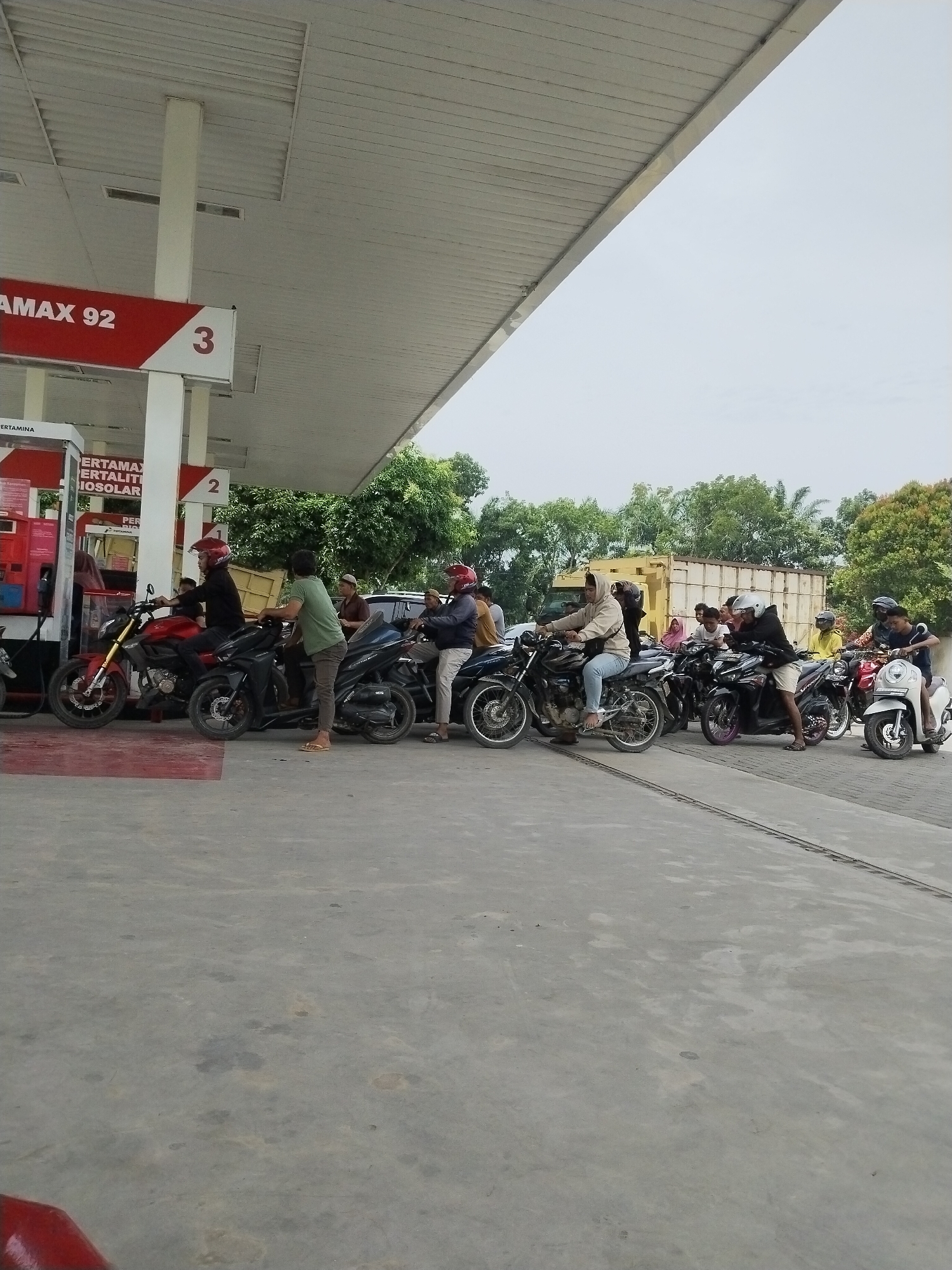 A line of motorcycles and their riders are waiting at a Pertamina gas station in Tanjung Putus, Indonesia. The gas station canopy extends over the waiting area, with pumps visible on the left. A red sign with "AMAX 92" is prominently displayed. In the background, trees and a yellow truck are visible. The ground is paved concrete, with some stains. The sky is overcast.