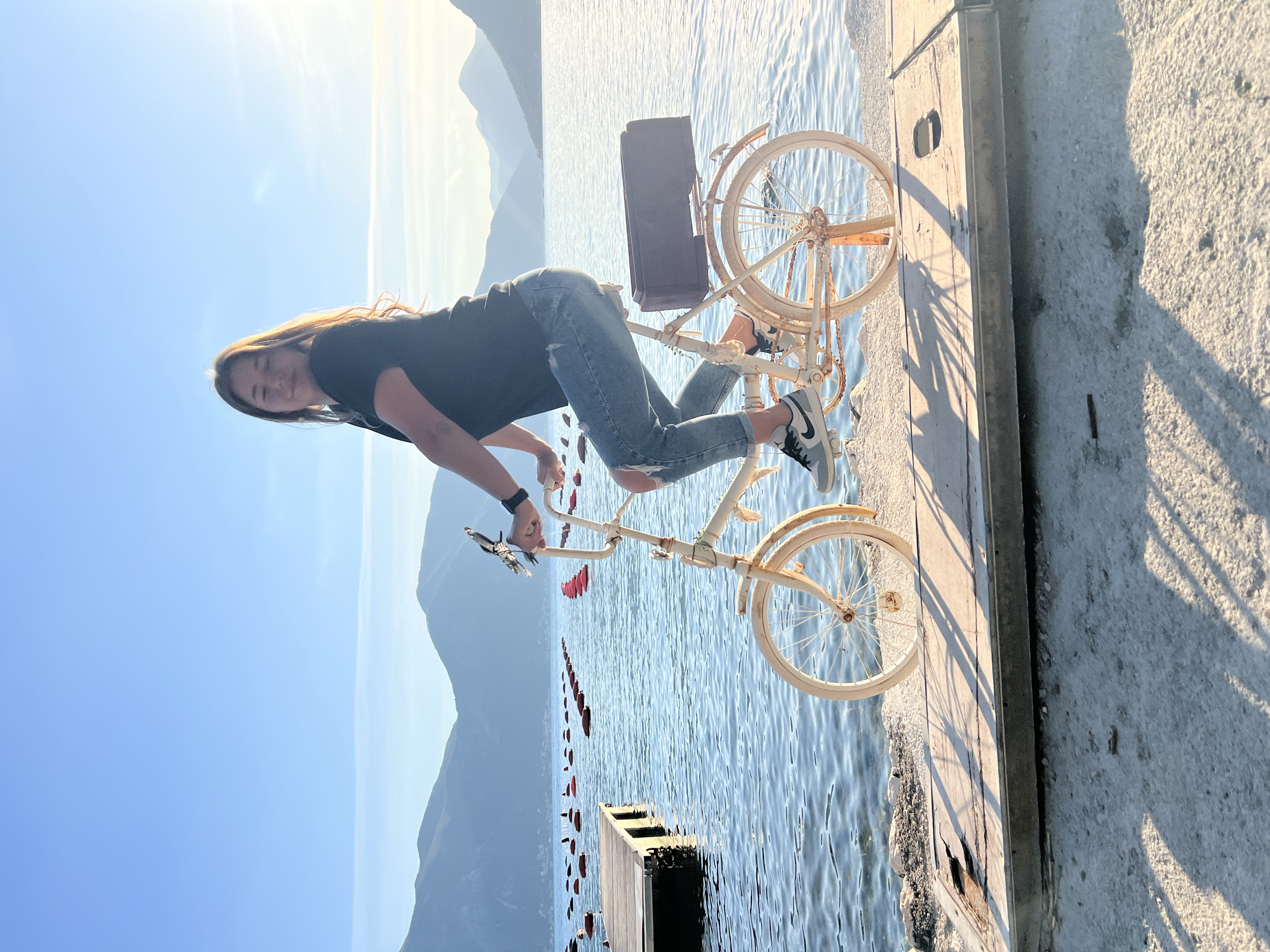 A person with long brown hair is leaning on a white, antique bicycle with a suitcase attached to the back, positioned on a wooden dock. The person is wearing a dark t-shirt, light blue jeans with rips, and grey and black sneakers. The bicycle faces the open water, where there are numerous red buoys and a submerged structure. In the background, mountains are visible under a clear, bright sky. The location is on the waterfront of Donji Orahovac, Montenegro.