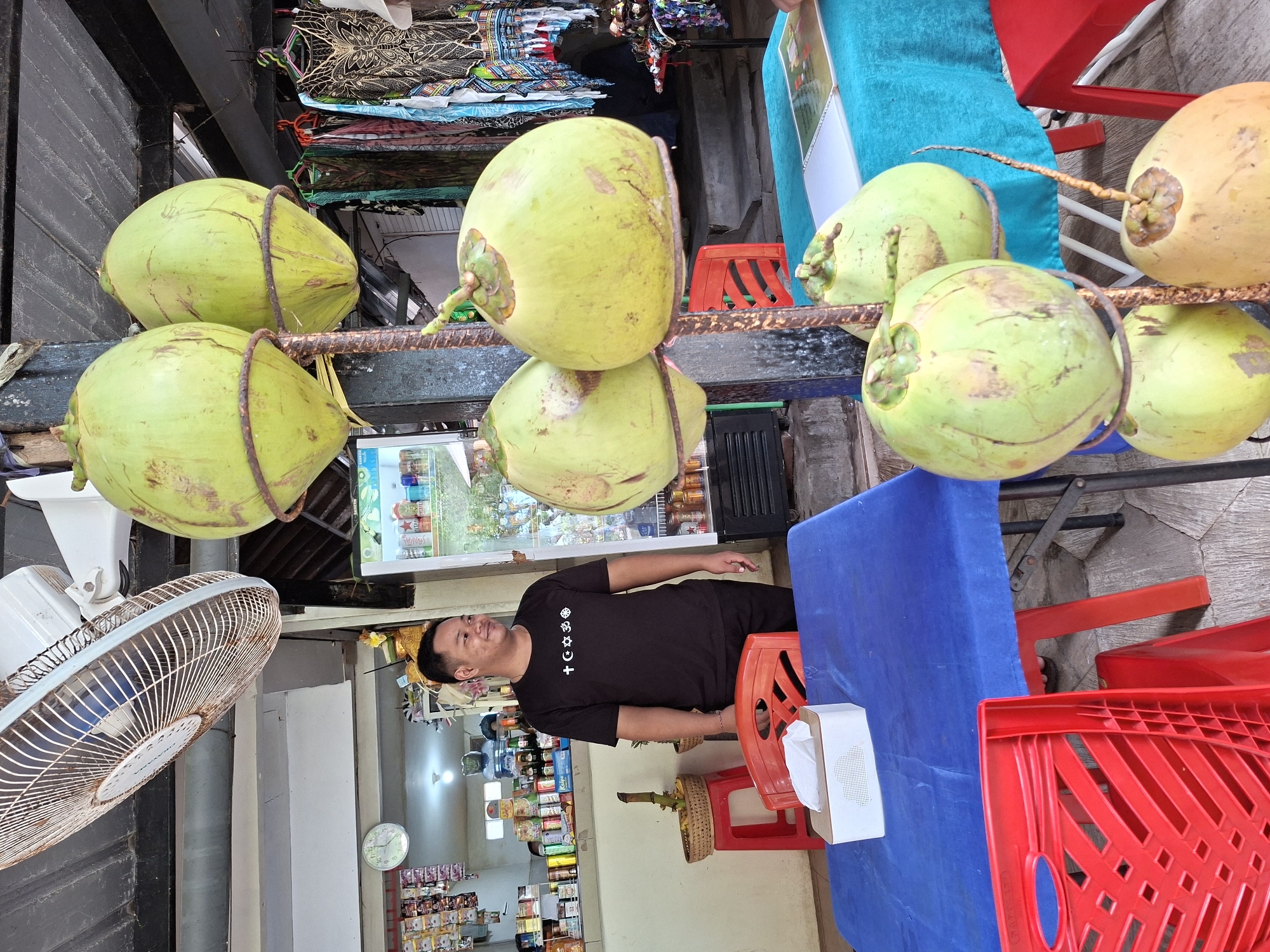 The image, rotated 90 degrees clockwise, presents a vibrant street-side stall scene in Kuta Selatan, Indonesia. The main subjects are a man and a prominent display of fresh coconuts, set against a backdrop of a small shop.
In the foreground, a row of approximately eight light green and yellowish-green coconuts are displayed, suspended from a rusty metal bar by ropes or resting directly on it. To the right, a blue-covered table is set with red plastic chairs, suggesting a customer seating area. A white tissue box sits on the table.
A man, presumably the vendor, stands to the left of the table. He has short, dark hair and wears a black t-shirt featuring a distinctive row of white symbols, including a cross within a circle, a star, a crescent moon, and an anchor. He is also wearing black shorts. He looks upward and to his right with a relaxed expression and a slight smile, seemingly at ease in his environment.
Behind the man, the stall's interior reveals a glass-front refrigerator stocked with various drinks, including cans labeled "Sarsi" and "STAR." Shelves laden with packaged snacks and goods line the back wall, where a white wall clock with green numerals is also visible. On the far left, a white oscillating fan is mounted, indicating warm conditions. Above the coconut display, neatly folded textiles or clothing items are stacked. The ground is paved with light-colored stones. The scene conveys a relaxed daytime atmosphere typical of a tropical market or street stall.